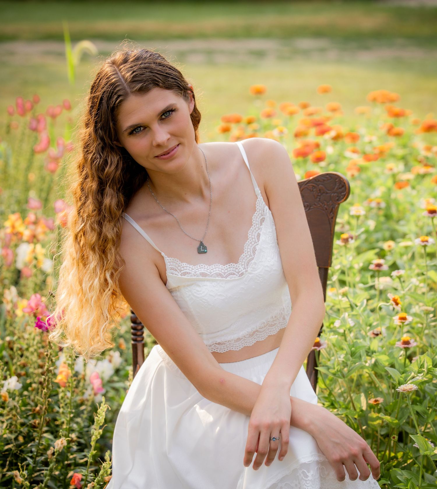 Young woman with curly hair in white crop top and skirt sits in a wooden chair surrounded by colorful wildflowers.
