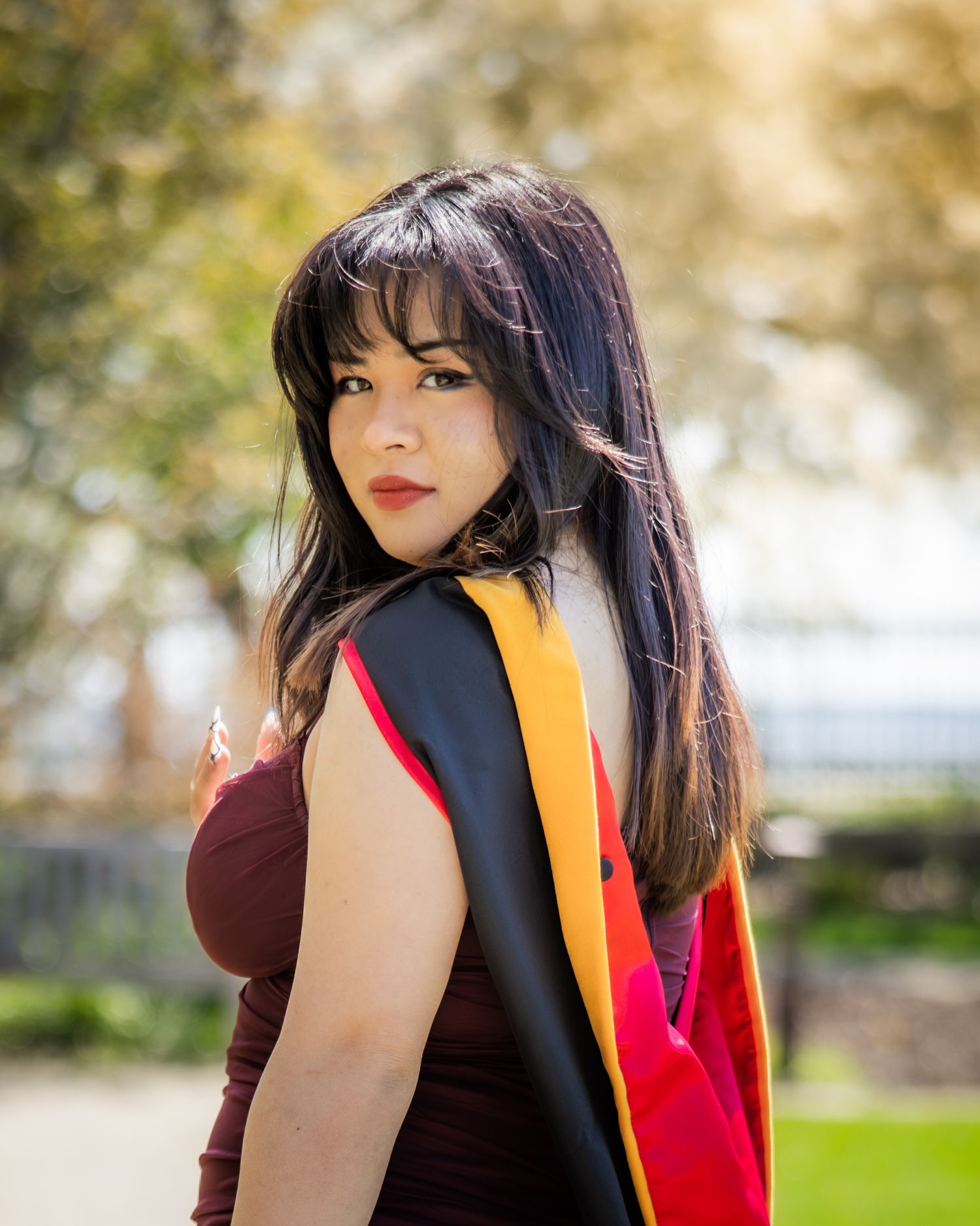 Young female graduate in maroon dress wearing black and red graduation sash posing outdoors in sunlight.
