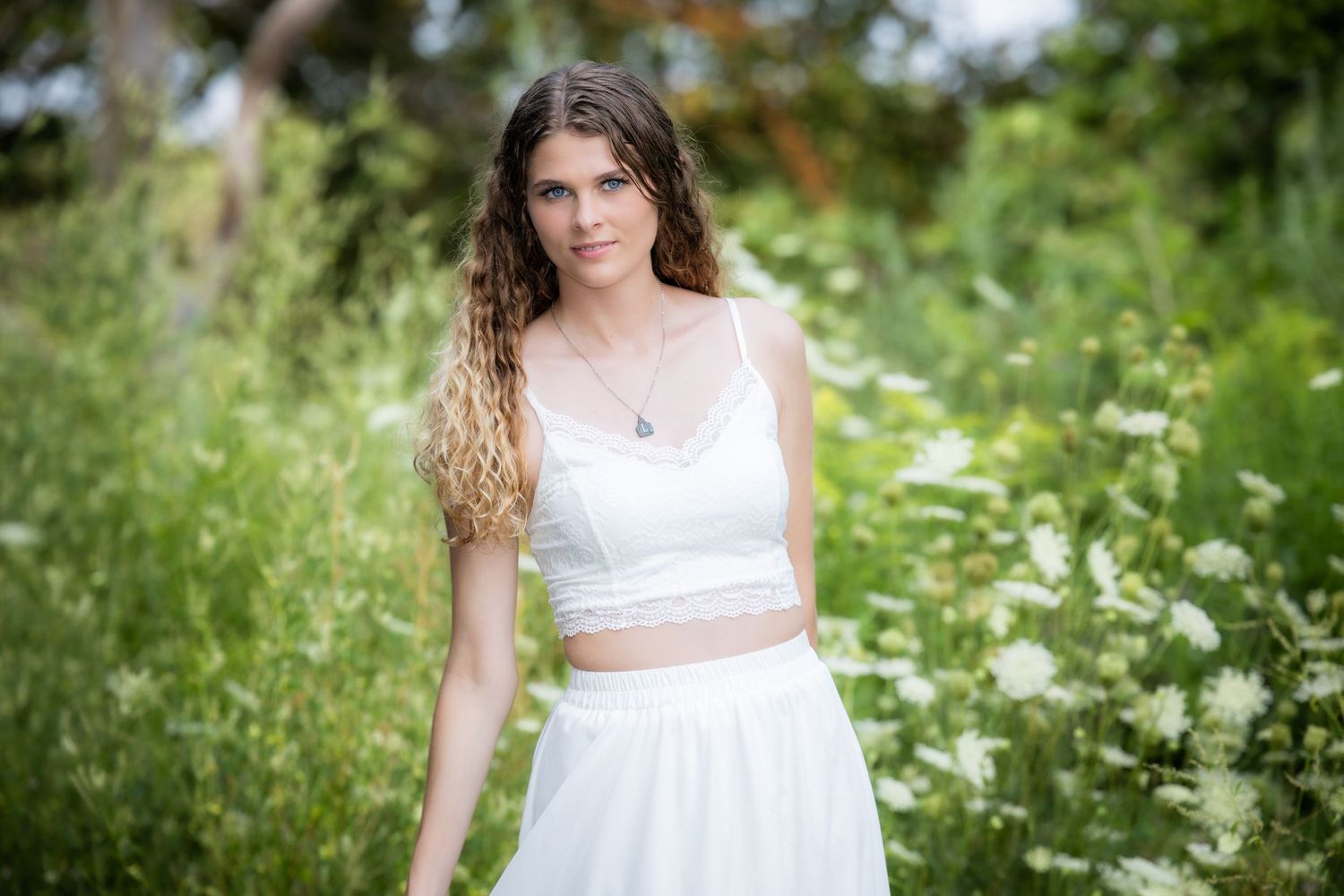 Young woman in white crop top and skirt poses outdoors among lush green foliage and wildflowers.