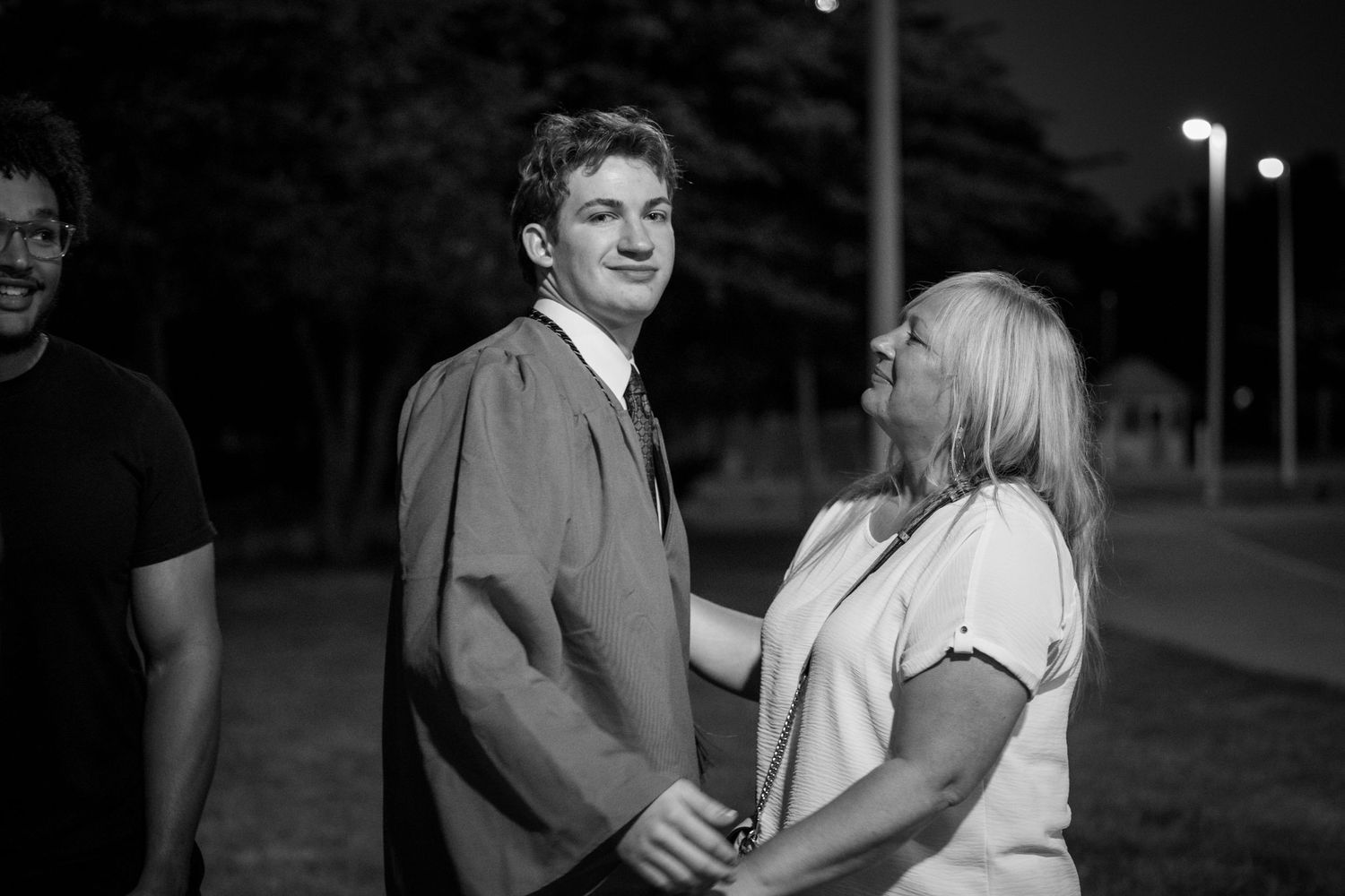 Young man in graduation gown smiling with woman outdoors at night.
