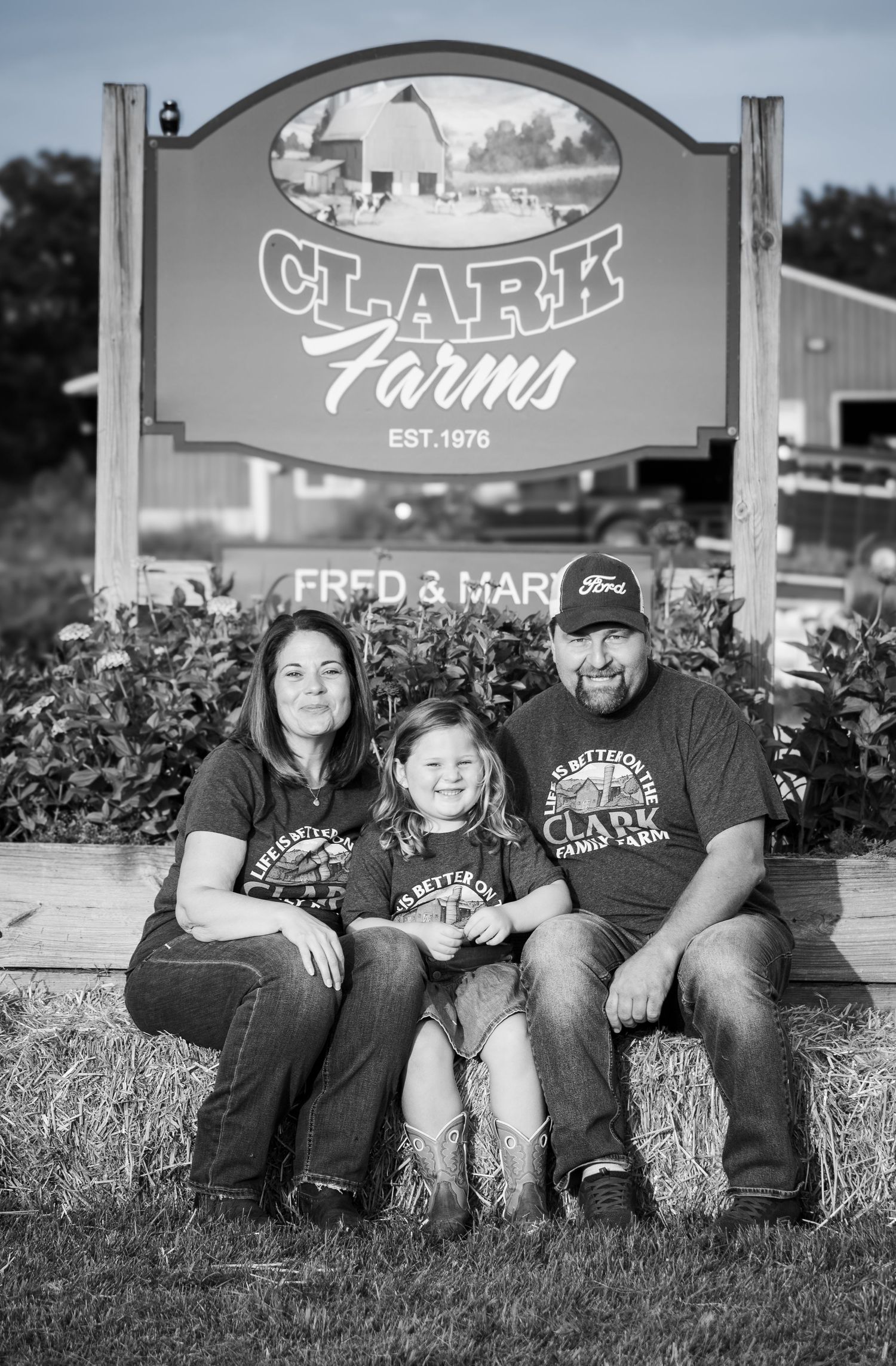 A couple and young child sit together in front of the Clark Farms Est. 1976 sign.