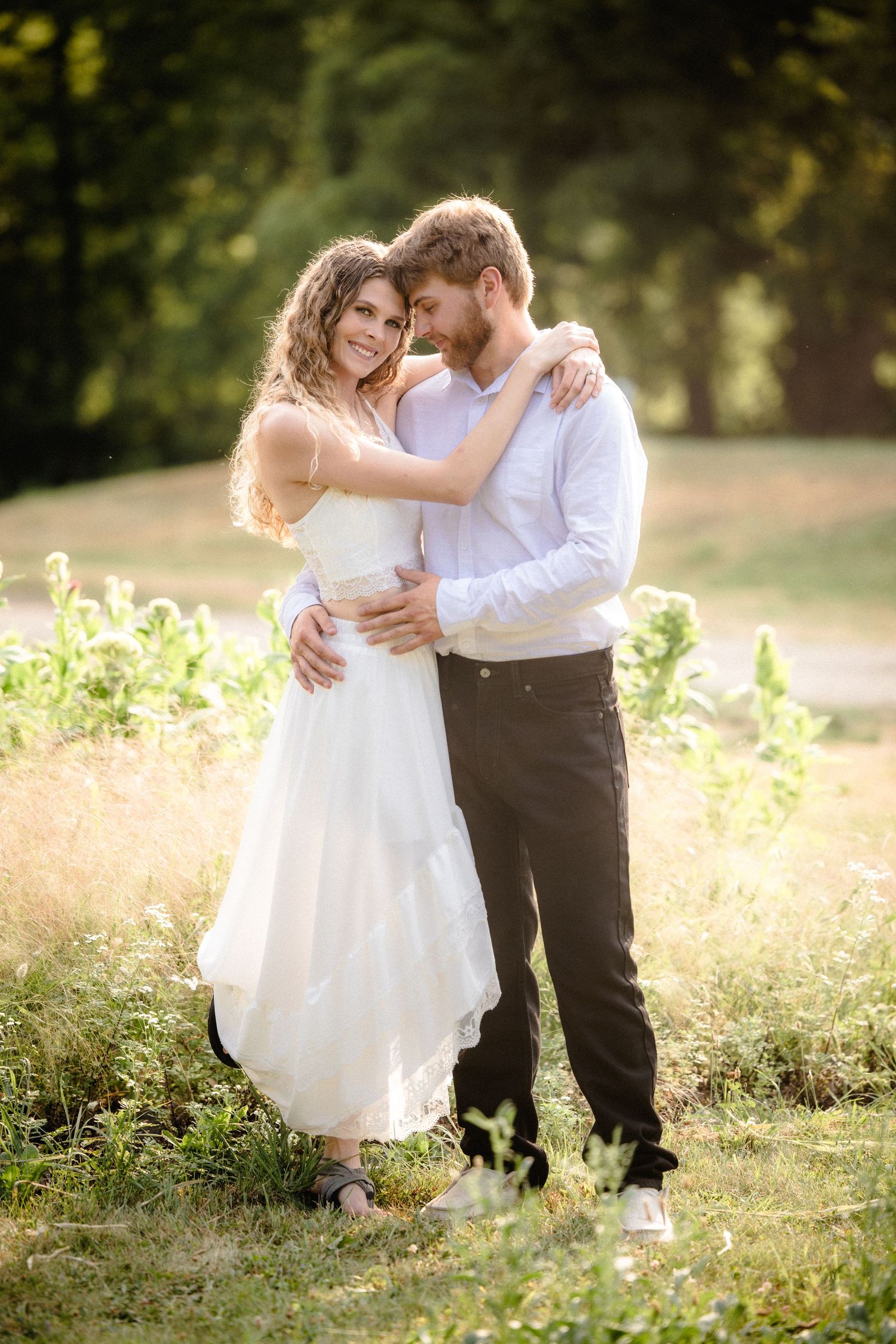 Couple embracing in sunlit park, bride in white dress and groom in white shirt, sharing a romantic outdoor moment.