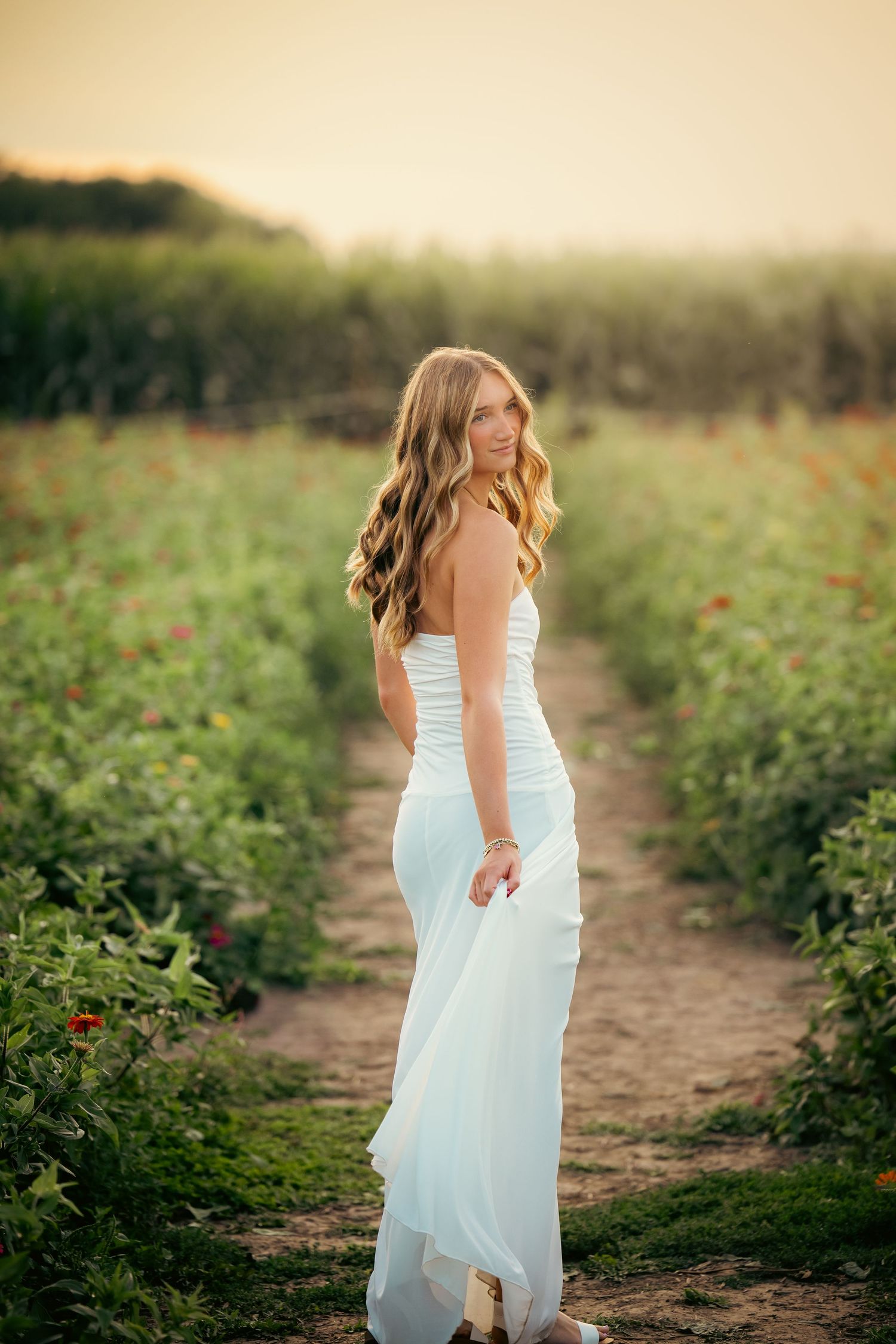 Young woman in white strapless gown standing in lush green field at golden hour, looking back over shoulder.