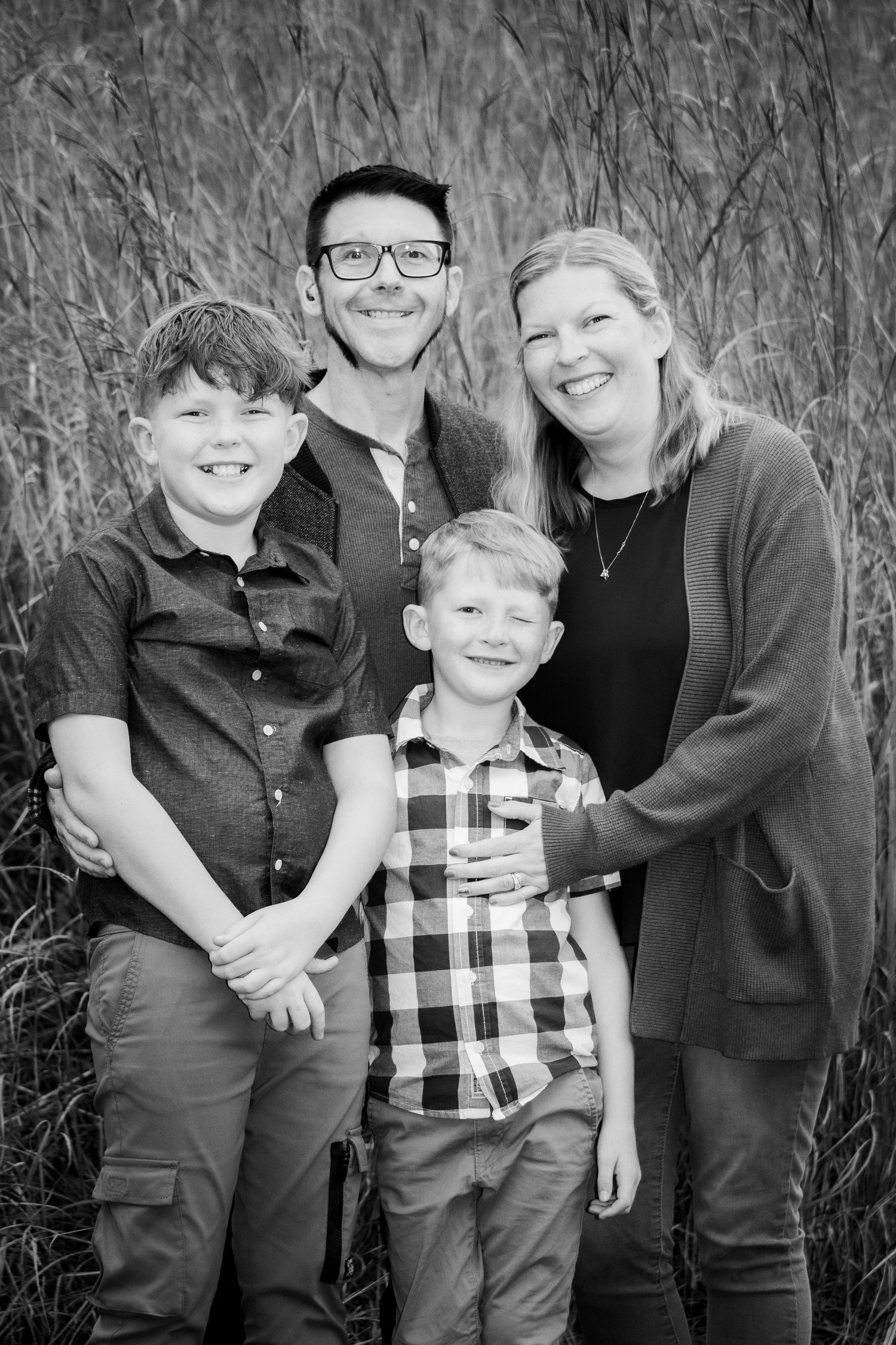 Black and white family portrait of smiling parents with two boys standing before tall grass.