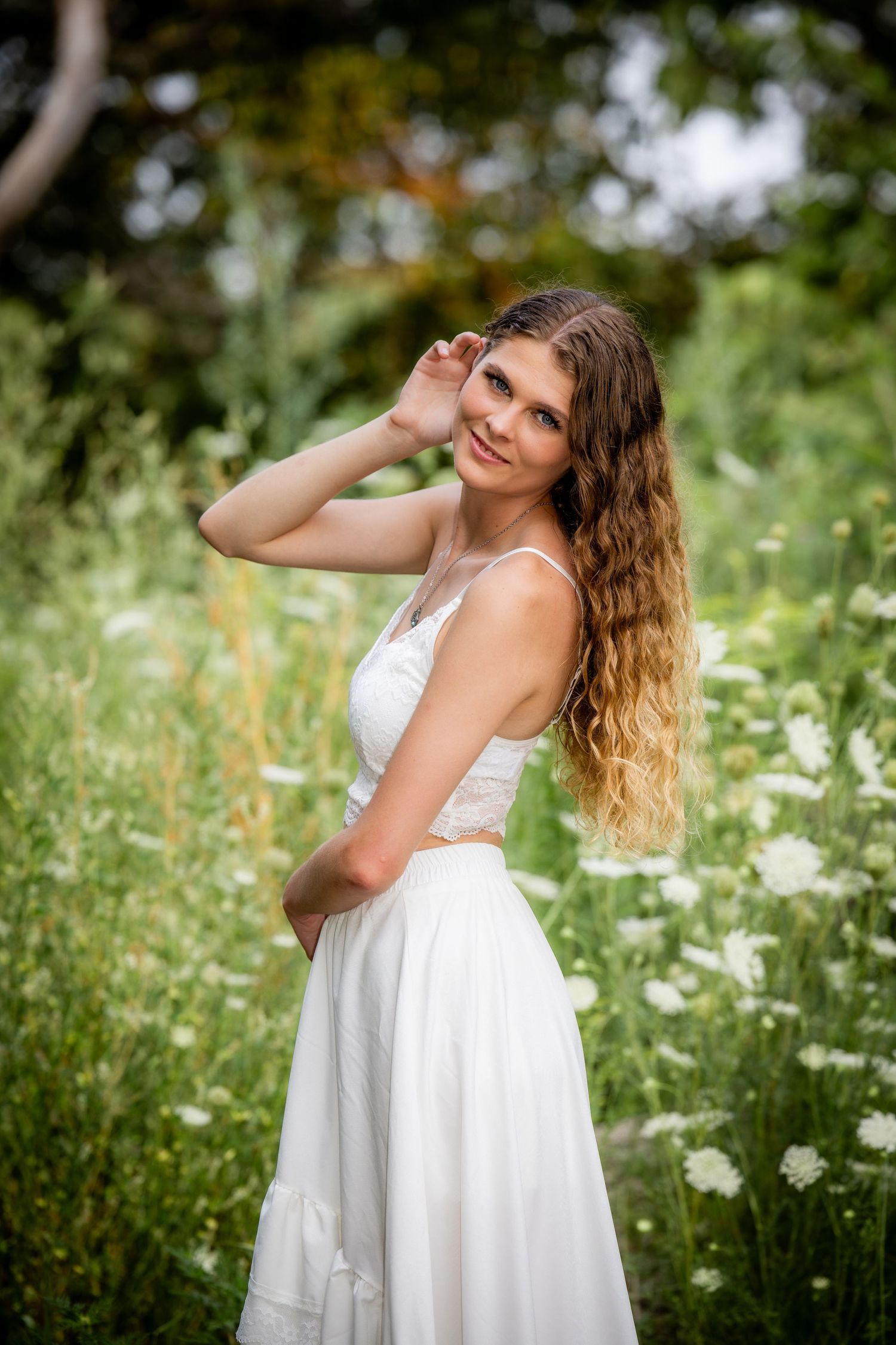 Young woman in white dress posing gracefully among wildflowers and daisies in a lush green meadow.