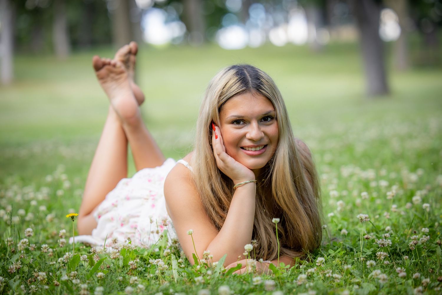 Senior in white dress lying on green grass field with wildflowers in a park.