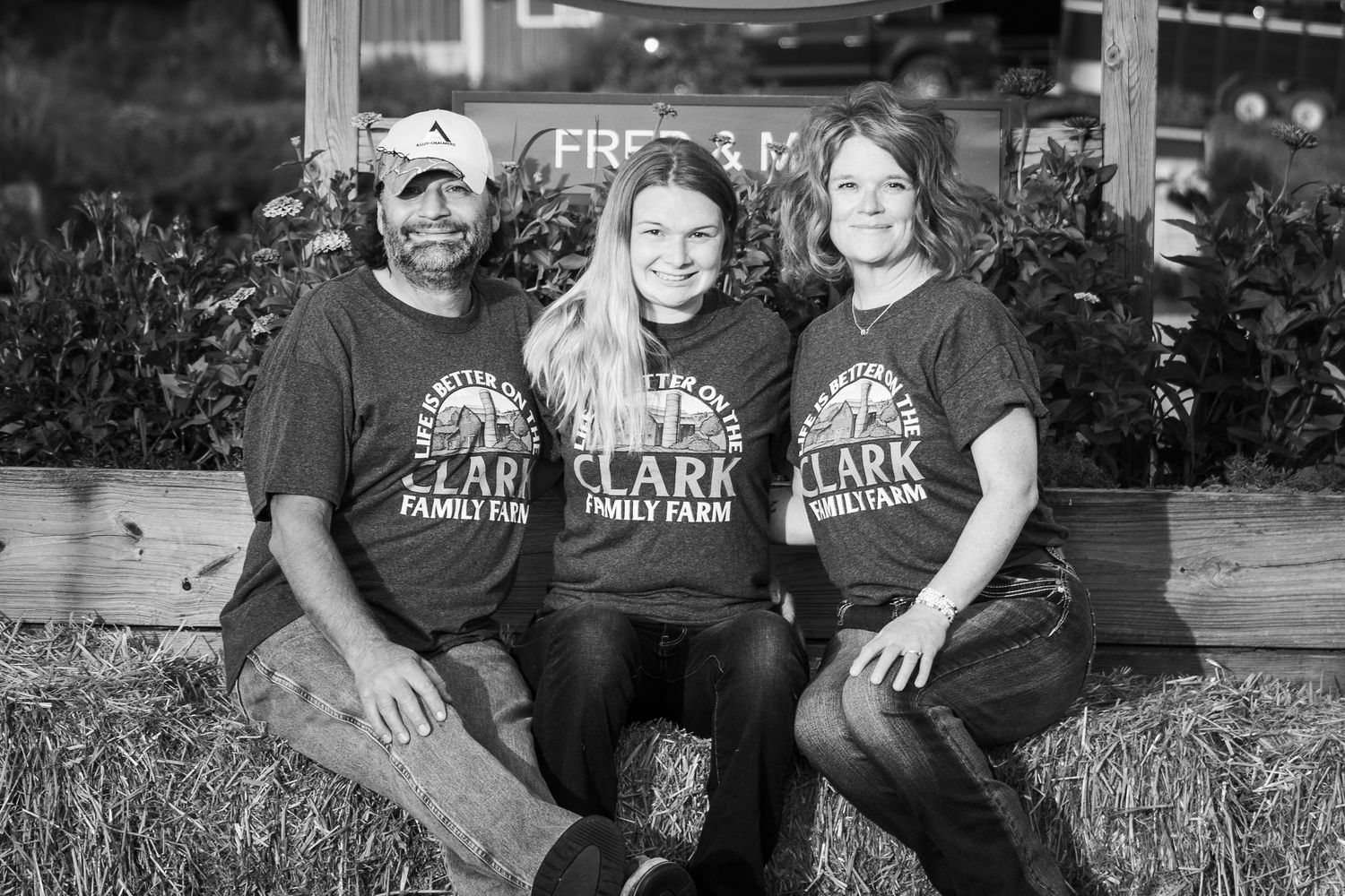 Three people in Clark Family Farm shirts sit together on hay bales in a warm outdoor photo.