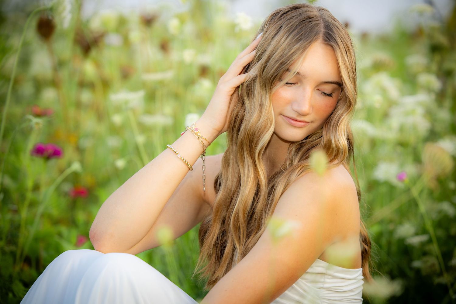 Young woman with long wavy blonde hair sits in a wildflower meadow, smiling softly with eyes closed.