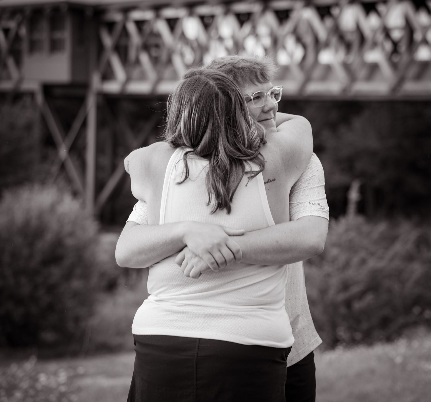 Two women share a warm embrace outdoors in black and white photo with a bridge in the background.