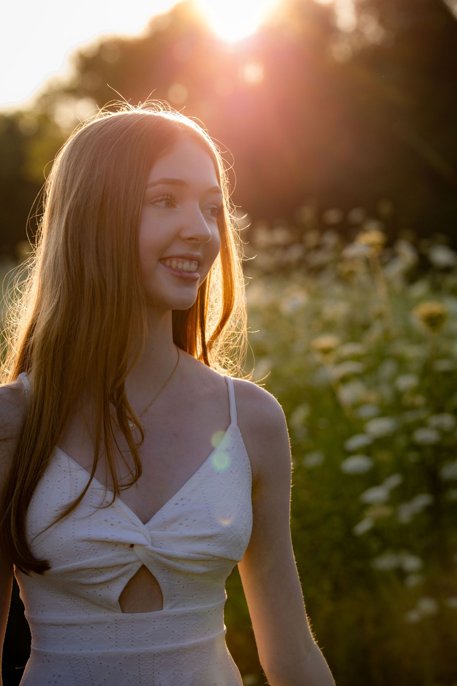 Young woman in white sundress smiling outdoors during golden hour sunset with wildflowers in background.