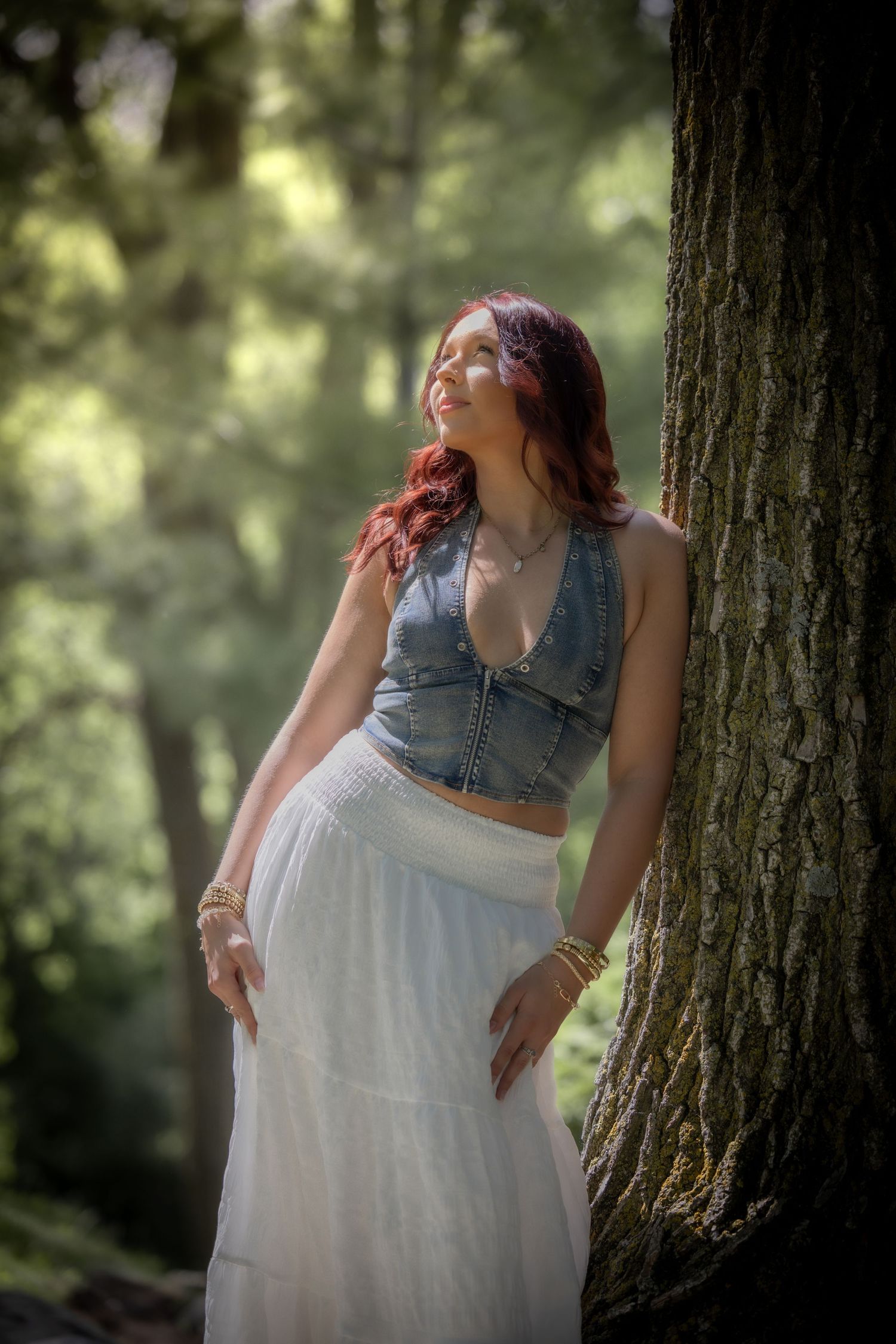 Senior in denim top and white skirt leaning against large tree in sunlit forest.