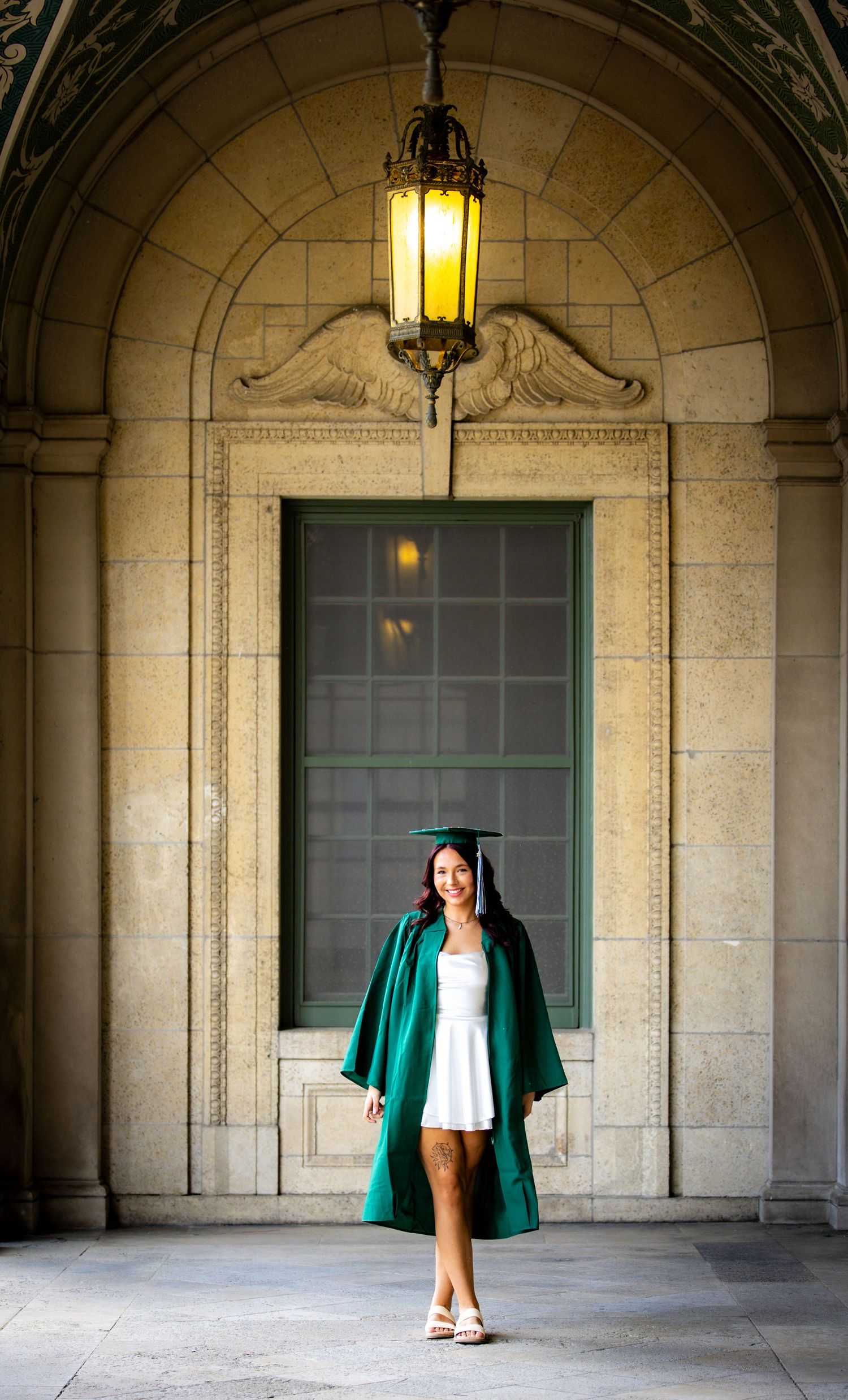 Female graduate in green cap and gown poses proudly under an elegant stone arch with glowing lantern.