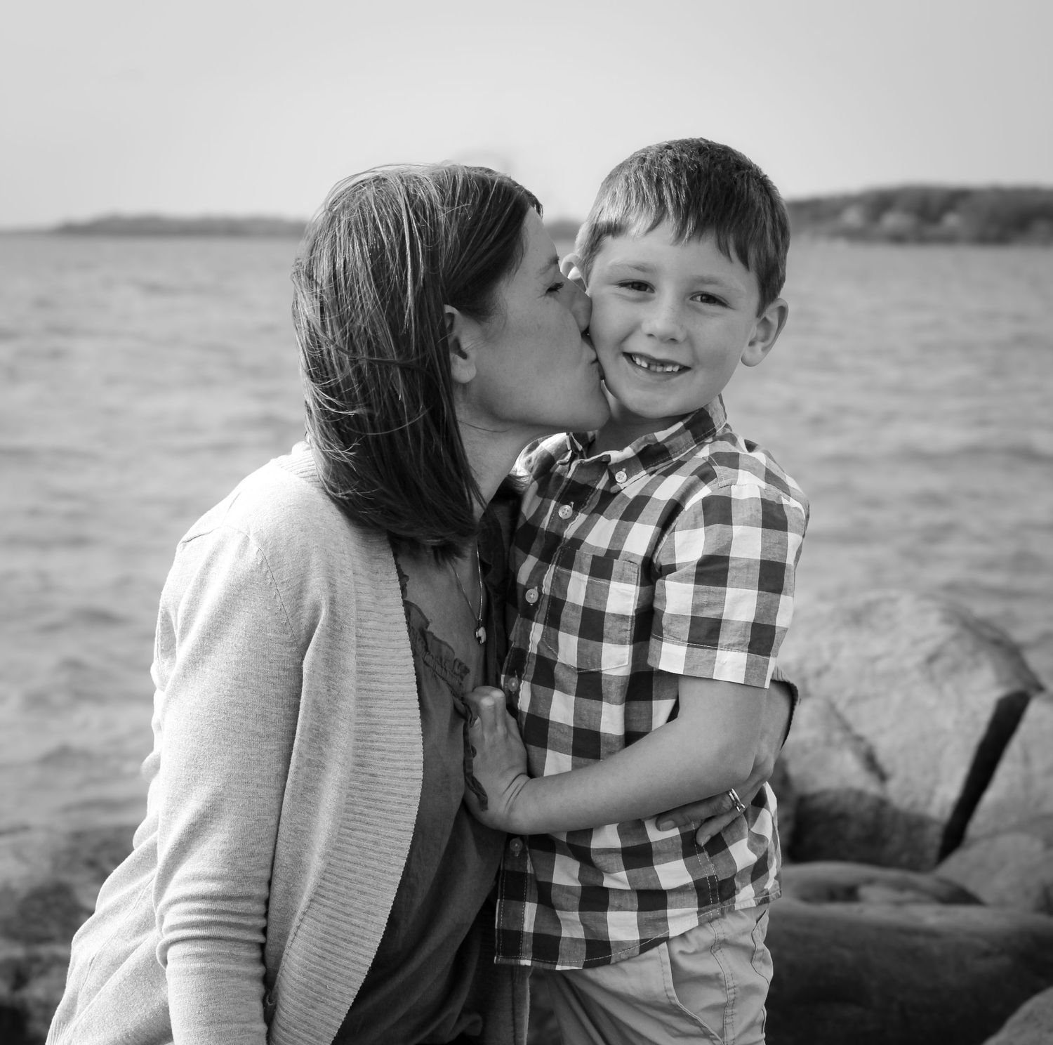 Mother kissing young smiling boy on cheek near rocky shoreline, black and white photo.