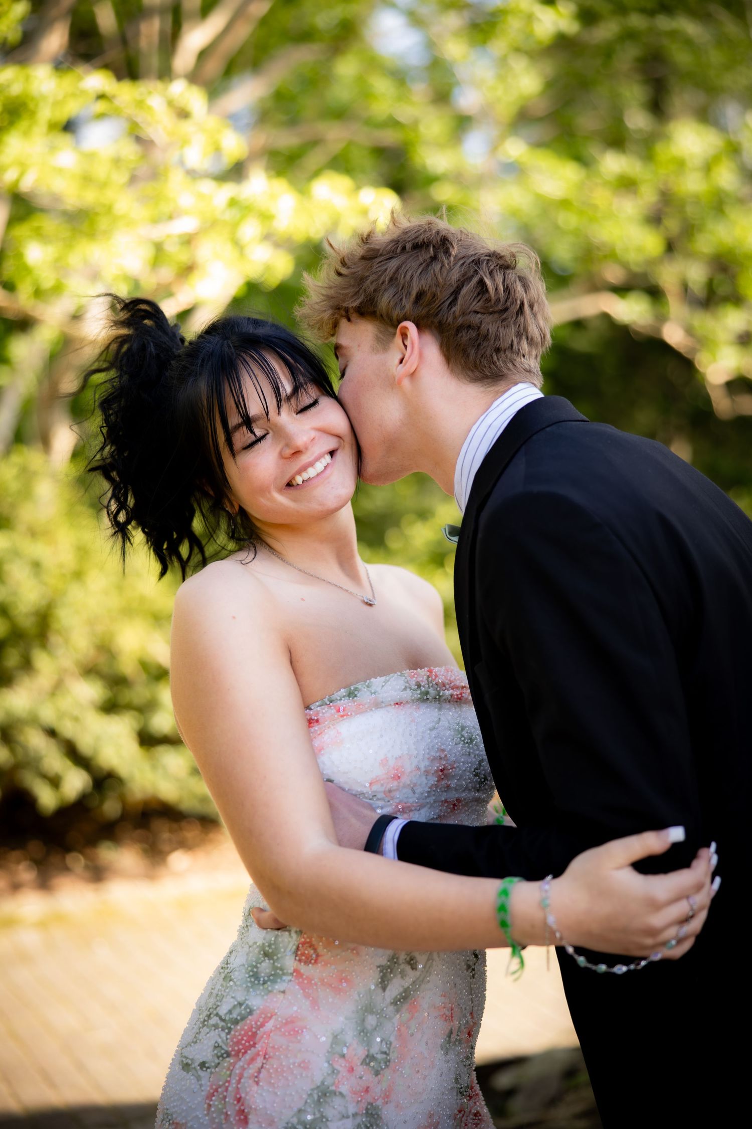 Young couple embracing outdoors, man kissing woman on cheek, both dressed for prom.