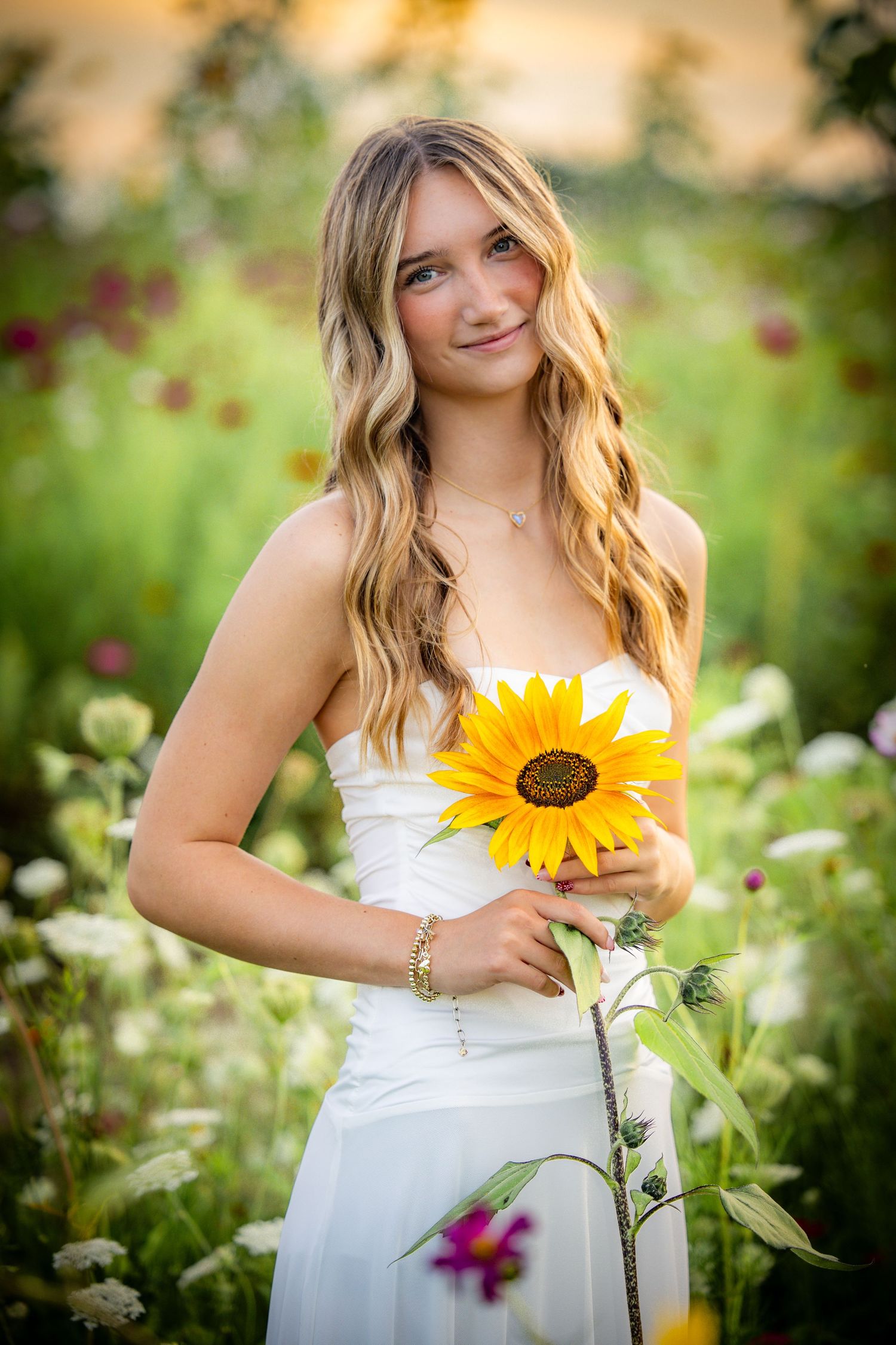 Young woman in white dress holding a sunflower, posing in a colorful wildflower field with soft golden light.