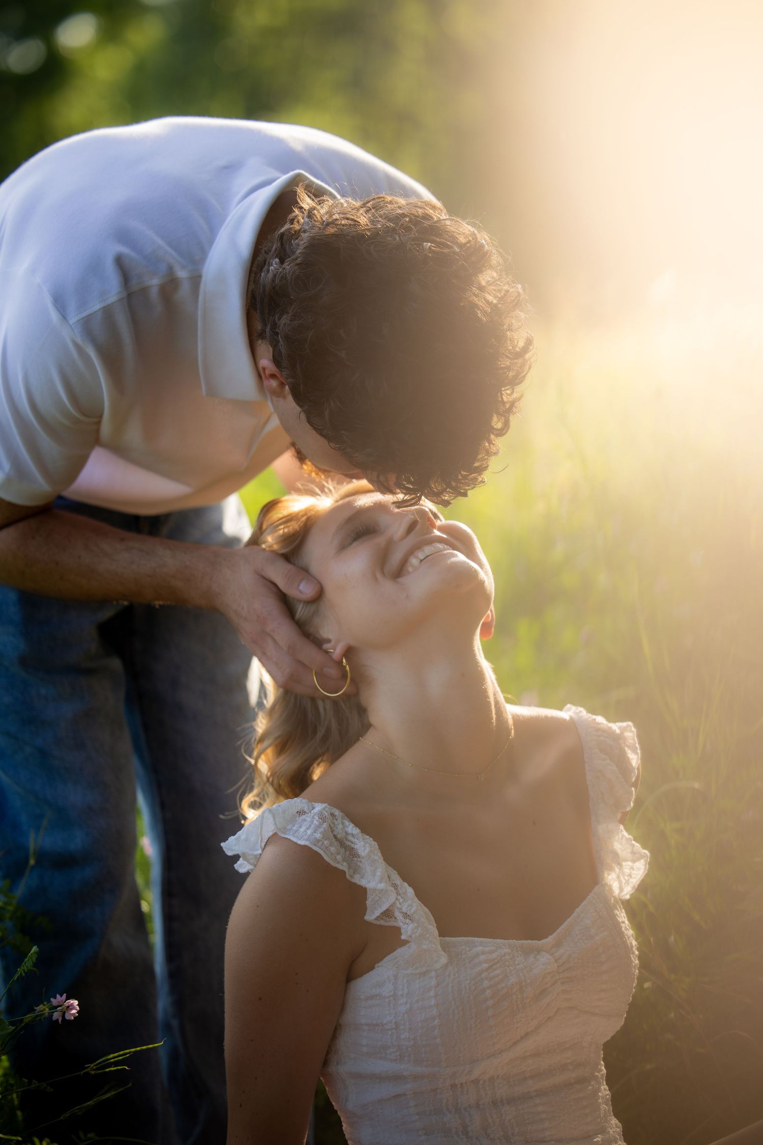 A young man gently kisses a smiling blonde woman on the forehead in golden sunlight outdoors.