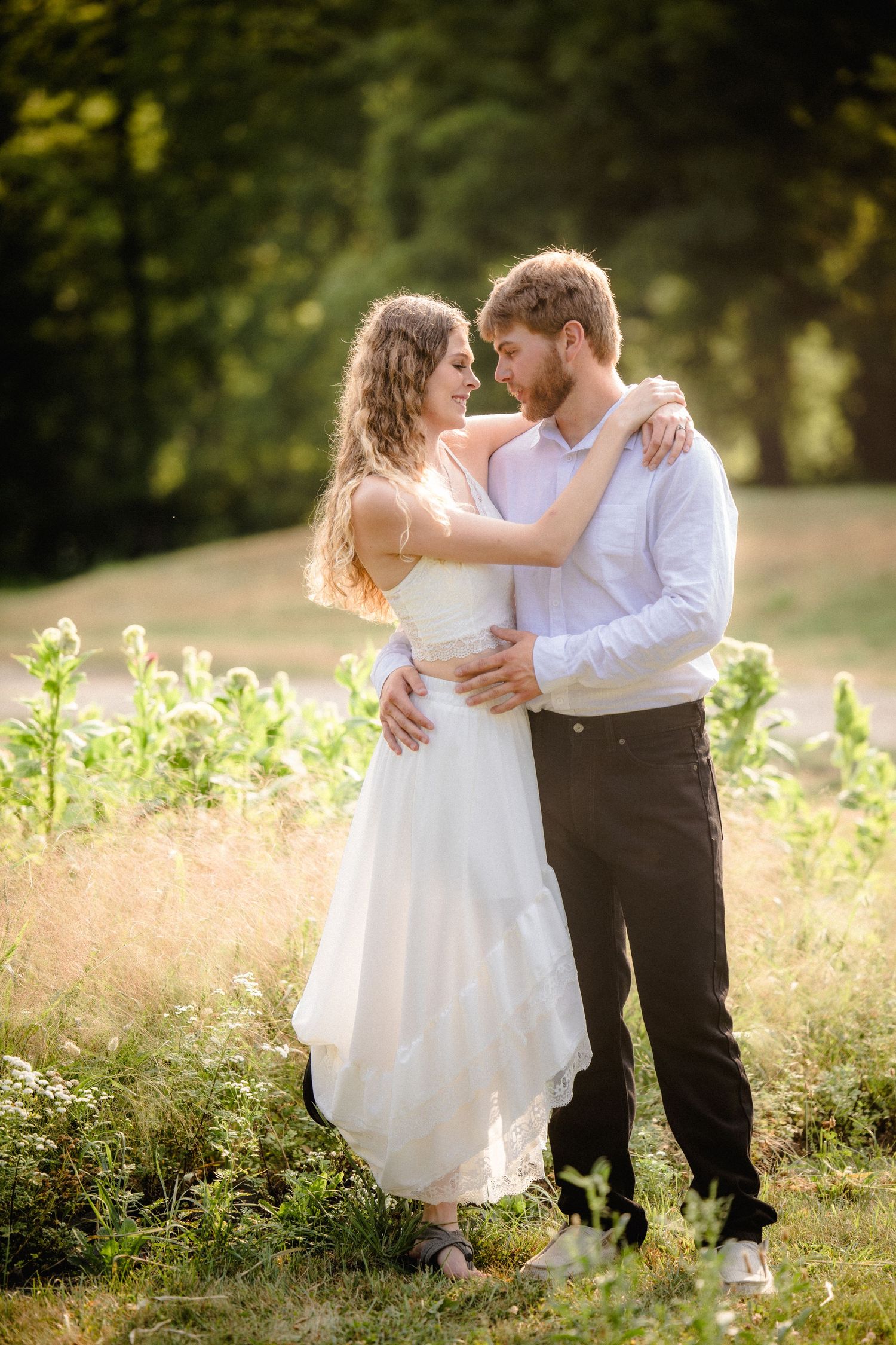 Couple in wedding attire embracing in a sunlit field, sharing a romantic moment surrounded by lush greenery.