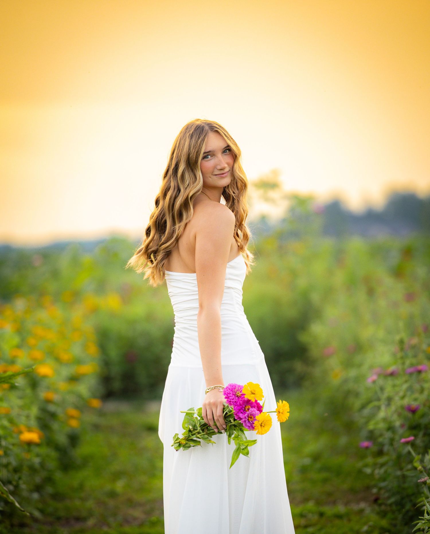 Young woman in white dress holding colorful flowers poses in a sunlit garden during golden hour.