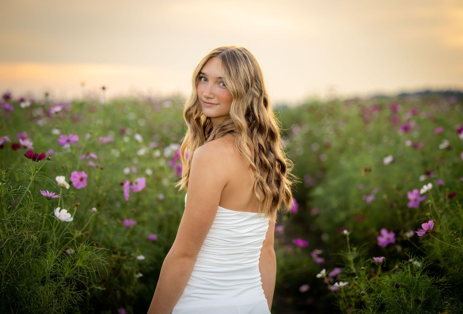 Young woman in white dress standing in a vibrant field of pink and purple cosmos flowers at golden hour.