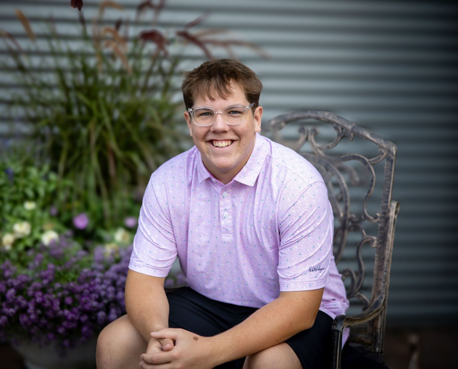 Smiling young man in pink polo shirt sitting on ornate chair outdoors with purple flowers and greenery behind him.