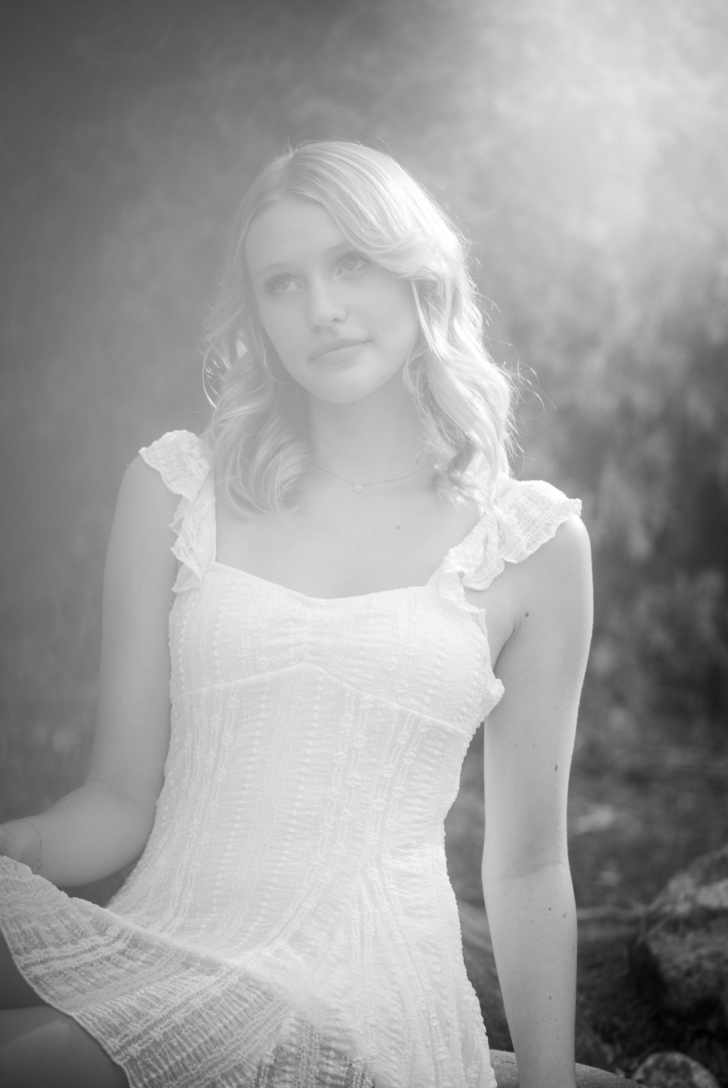 A black and white portrait of a blonde woman in a white dress sitting in soft dreamy light.