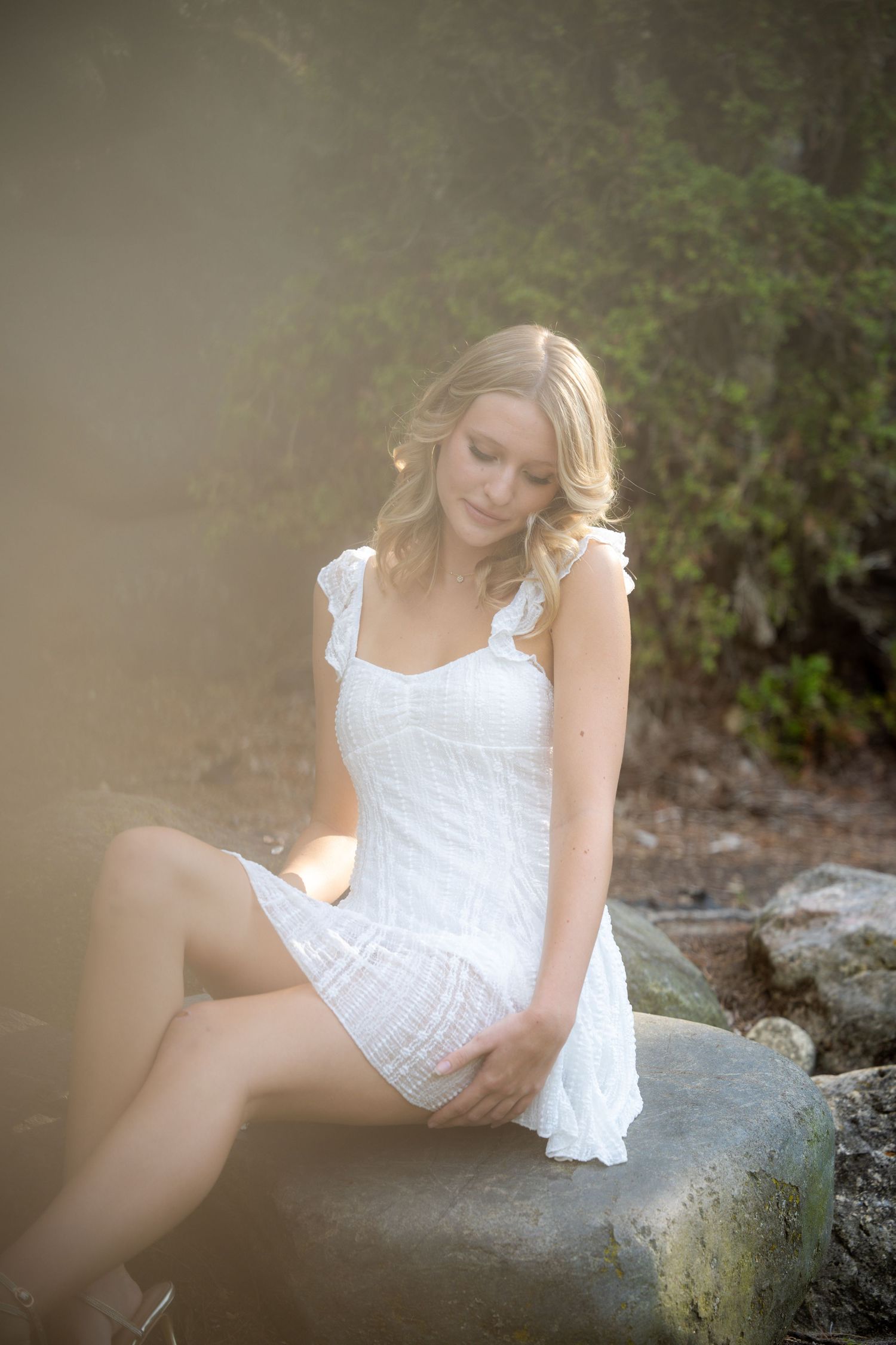 Young blonde woman in white sundress sitting on rocks outdoors, surrounded by soft misty light and green foliage.