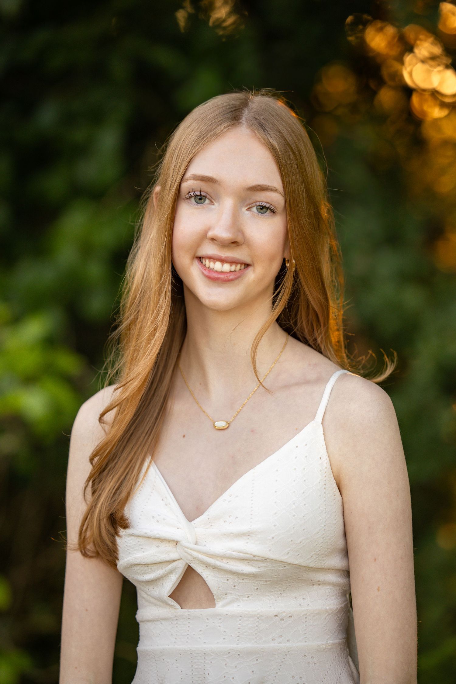 Young woman with long auburn hair smiling outdoors in white dress with gold necklace, bokeh green background.