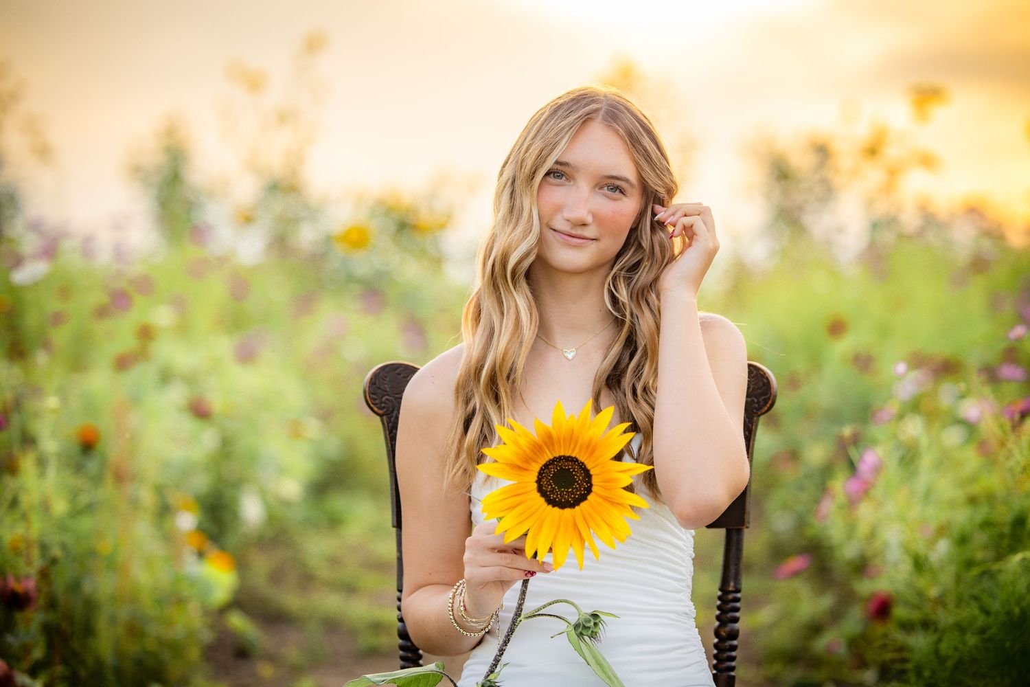 Young woman in white dress holding a sunflower, seated in a colorful flower field during golden hour.