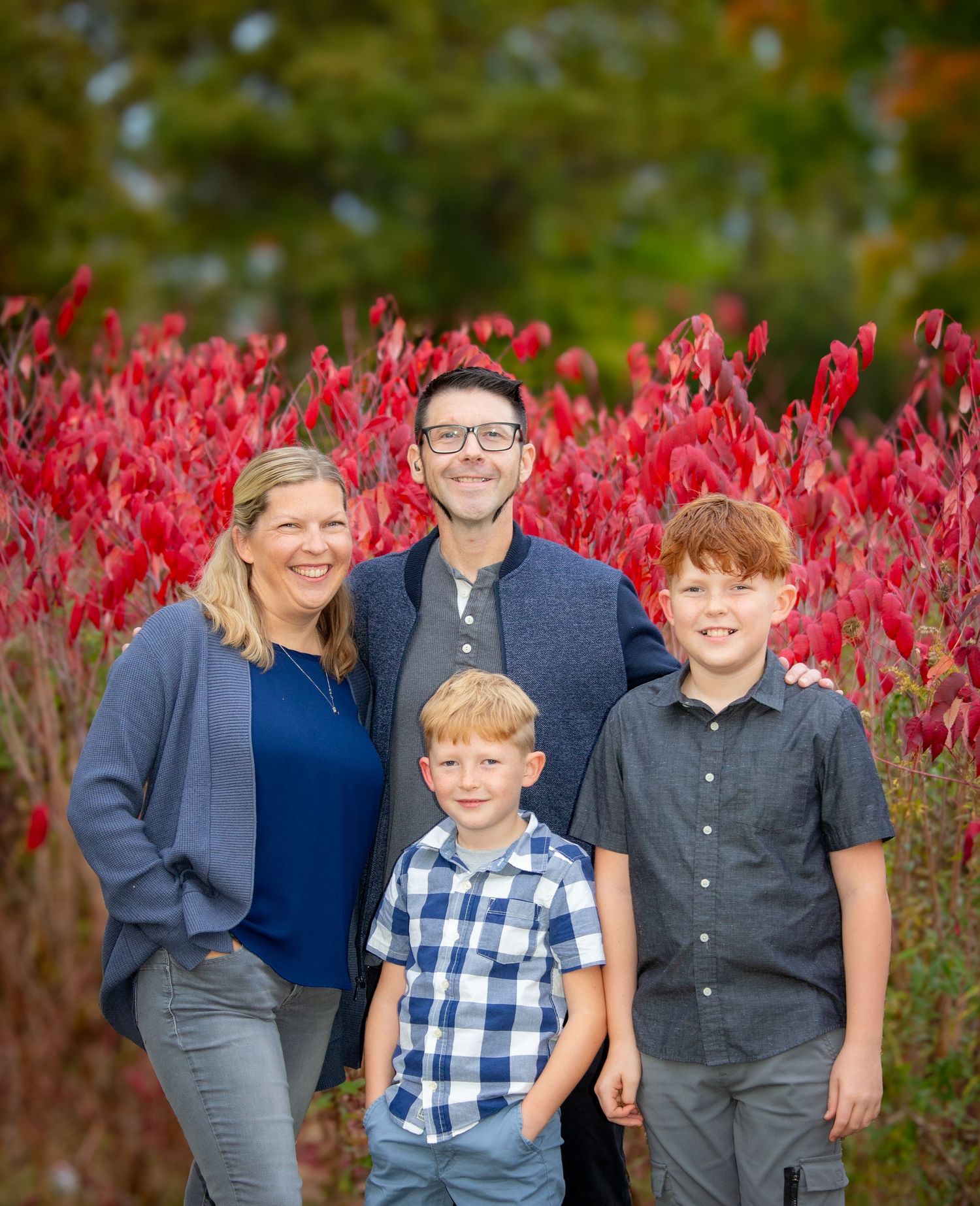 Happy family of four posing in front of vibrant red autumn foliage outdoors.
