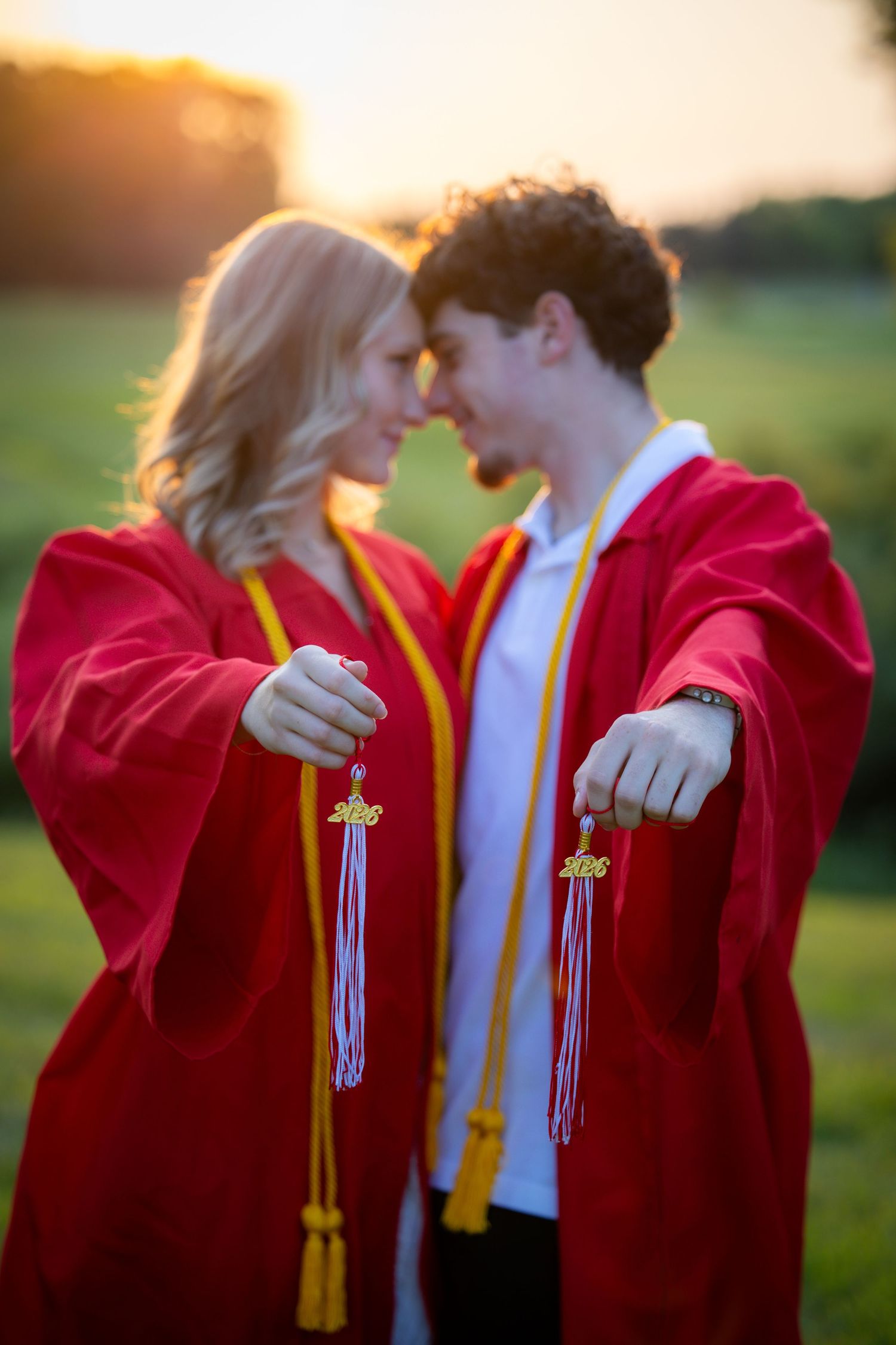 Two graduates in red robes holding 2020 graduation tassels facing each other at sunset.