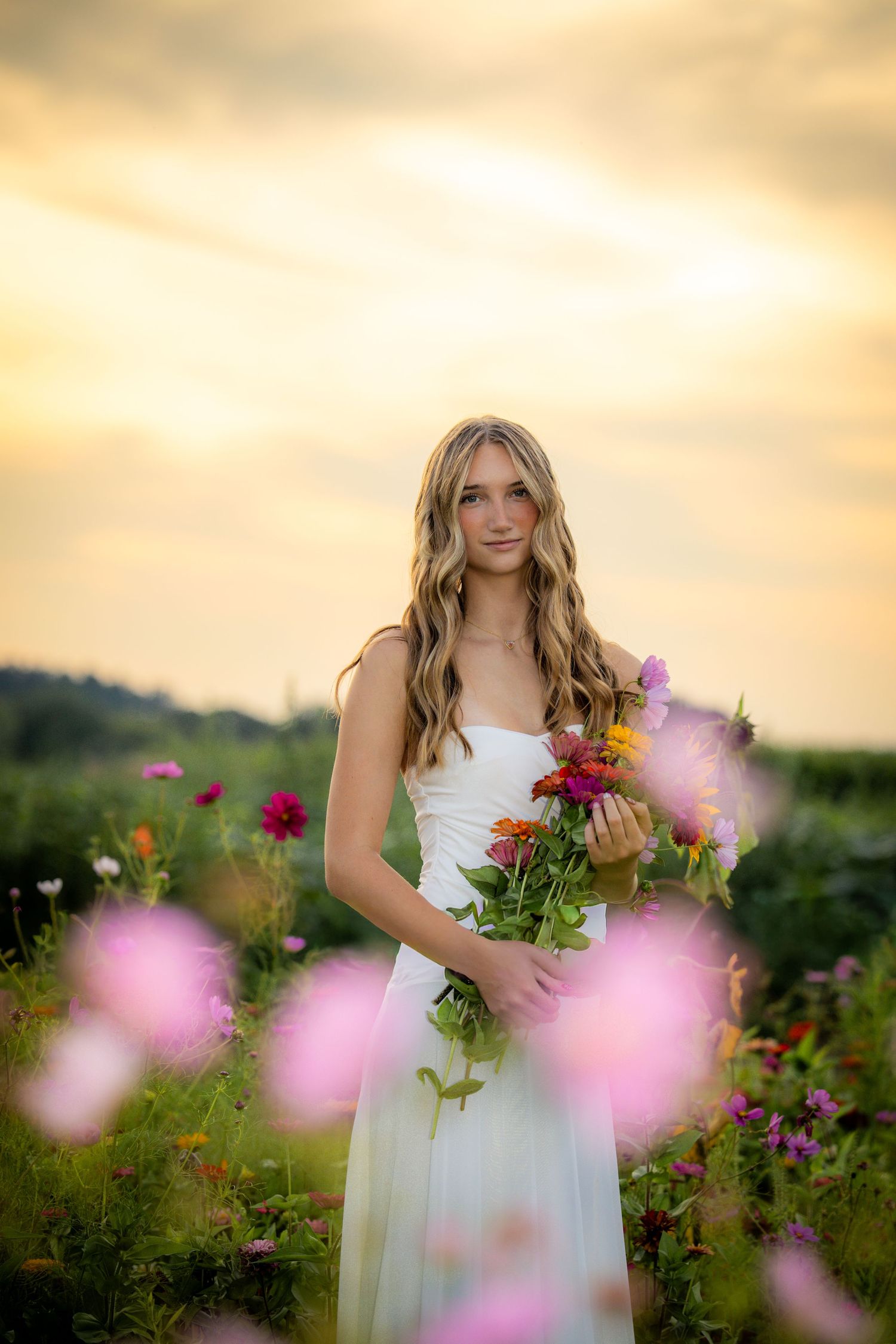 Young woman in white dress holding colorful bouquet in flower field at golden sunset.
