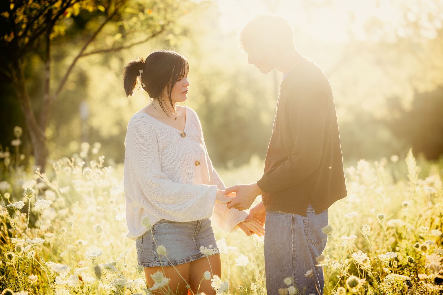 Couple holding hands in a sunlit wildflower meadow, golden backlight creating a romantic outdoor portrait.