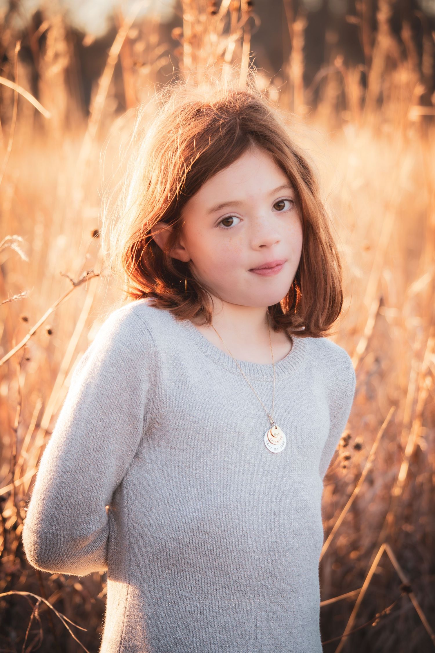 Young girl with shoulder length hair wearing gray sweater and necklace in golden sunlit field.