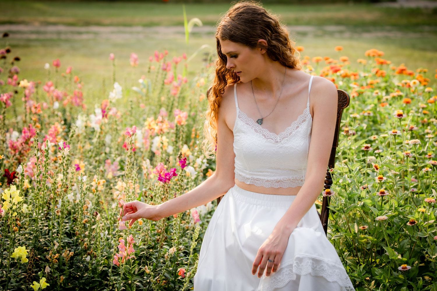 Young woman in white crop top and skirt sitting among colorful wildflowers in a sunny garden field.