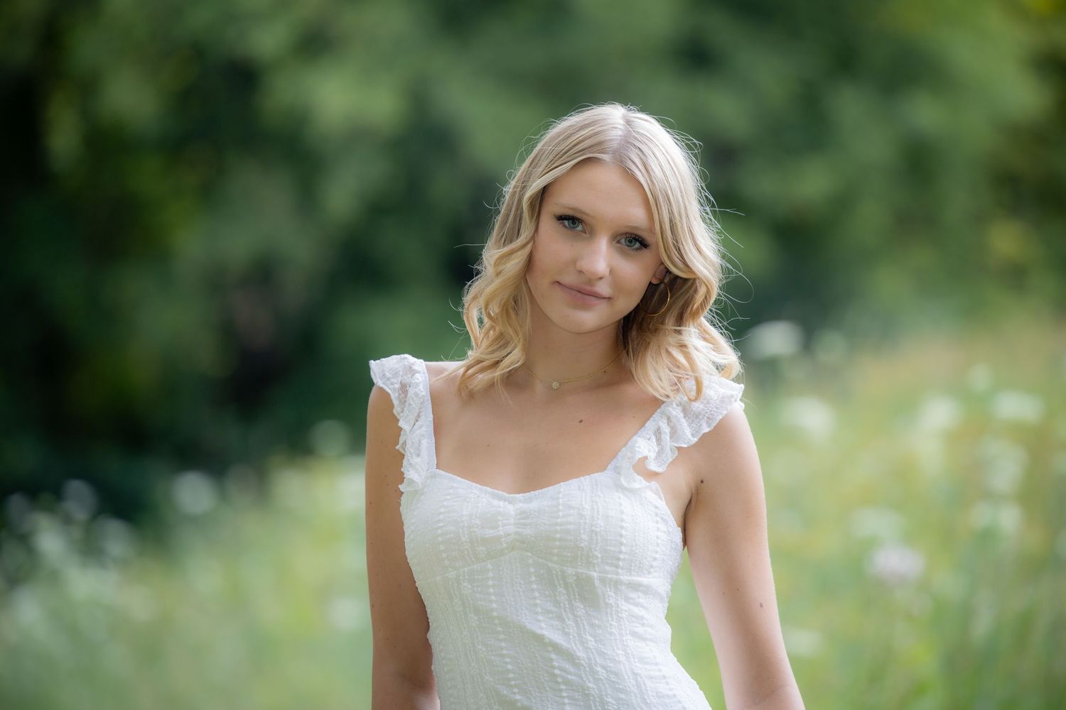 A smiling blonde woman in a white ruffled dress stands in a lush green wildflower meadow.
