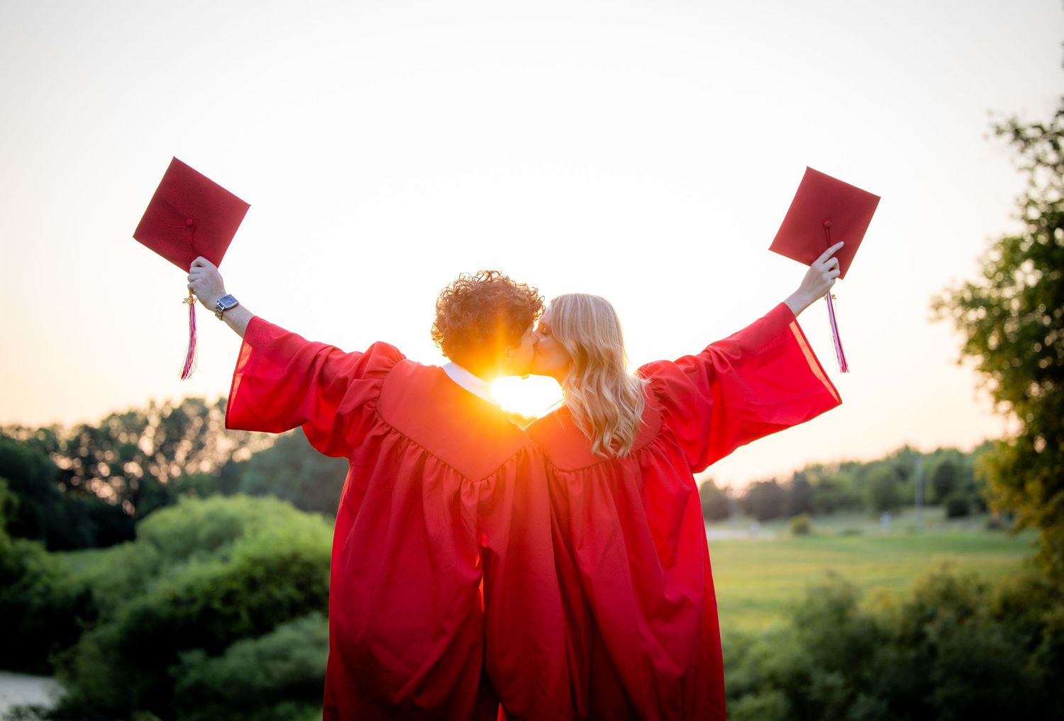 Two graduates in red caps and gowns kissing while raising mortarboards at golden sunset outdoors.