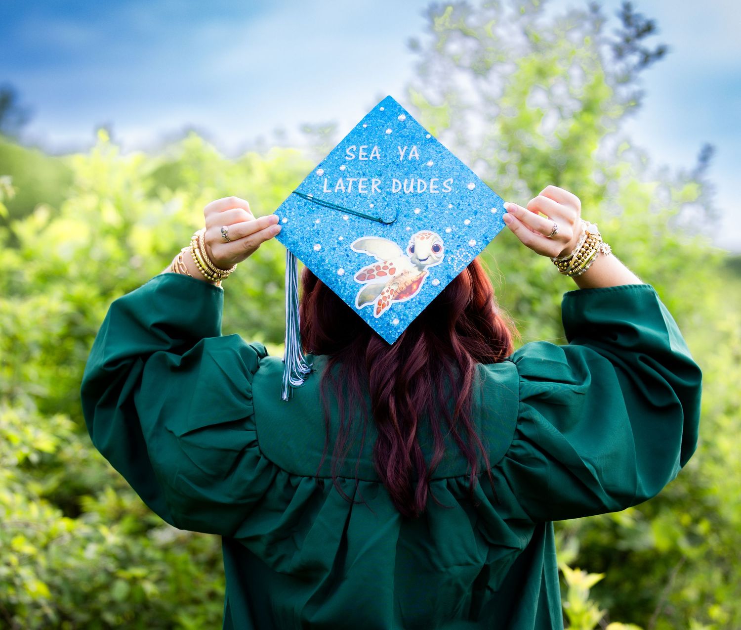 Graduate in green gown displays decorated cap reading Sea Ya Later Dudes with ocean art outdoors.