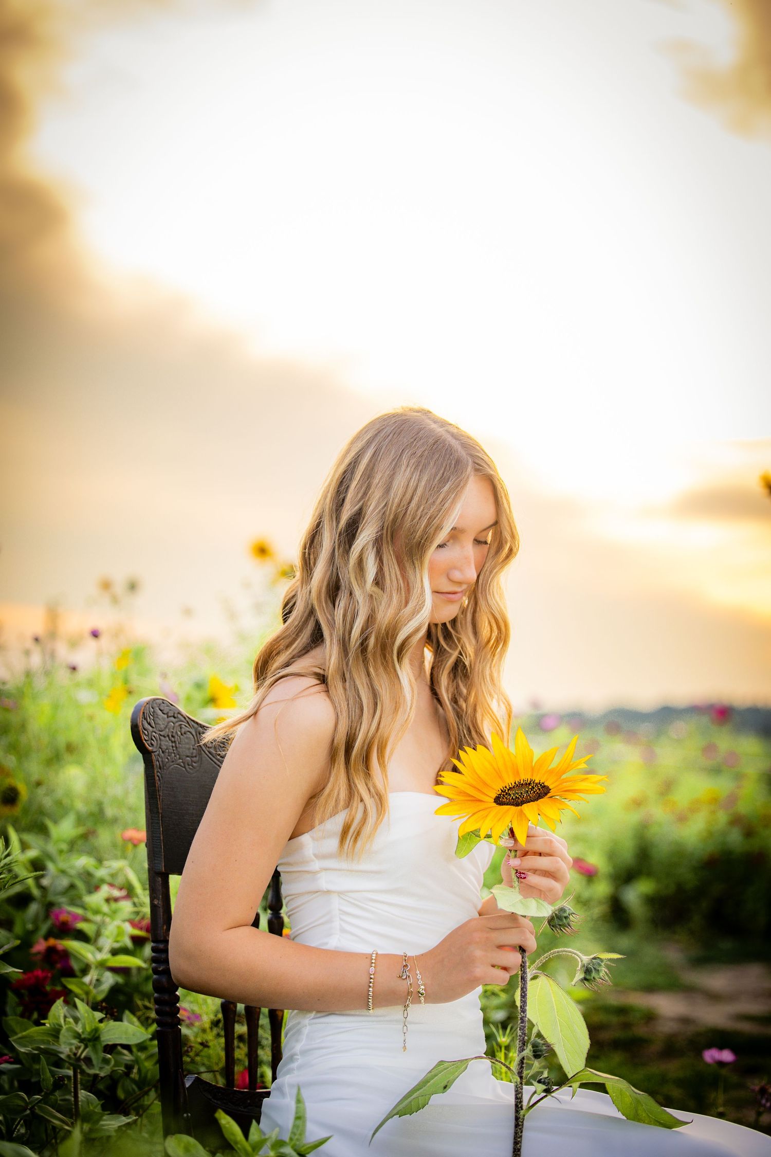 Young blonde woman in white dress holds a sunflower while sitting in a colorful flower field at golden sunset.