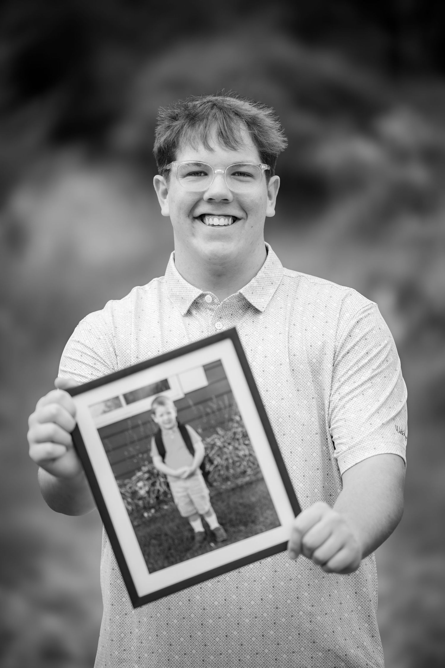 Smiling young man holds a framed childhood photo outdoors in a black and white senior portrait.