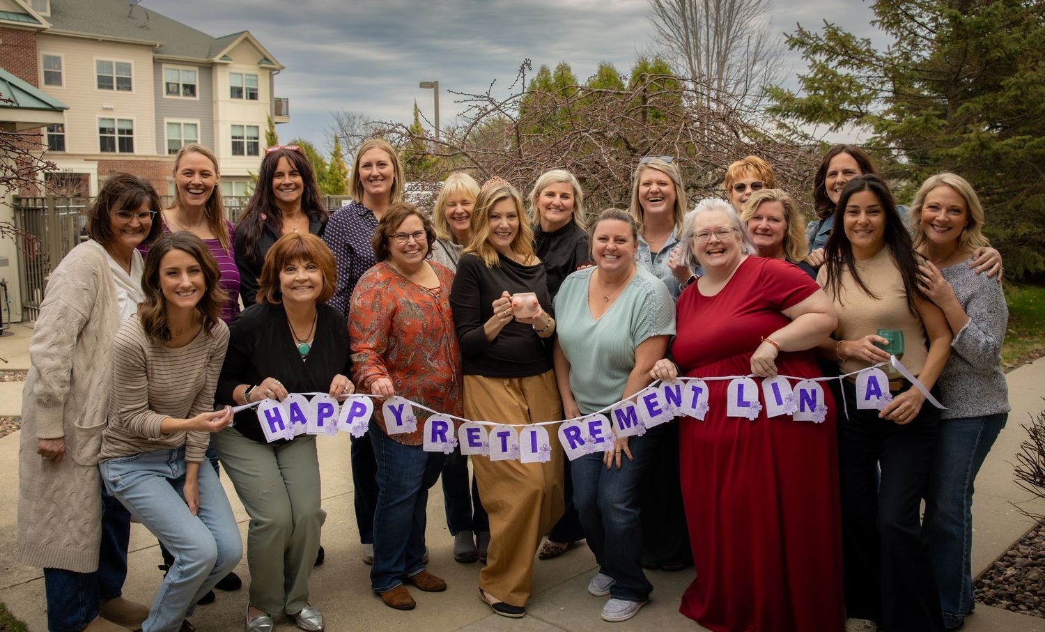 Group of women smiling outdoors holding a Happy Retirement Linda banner in a suburban setting.