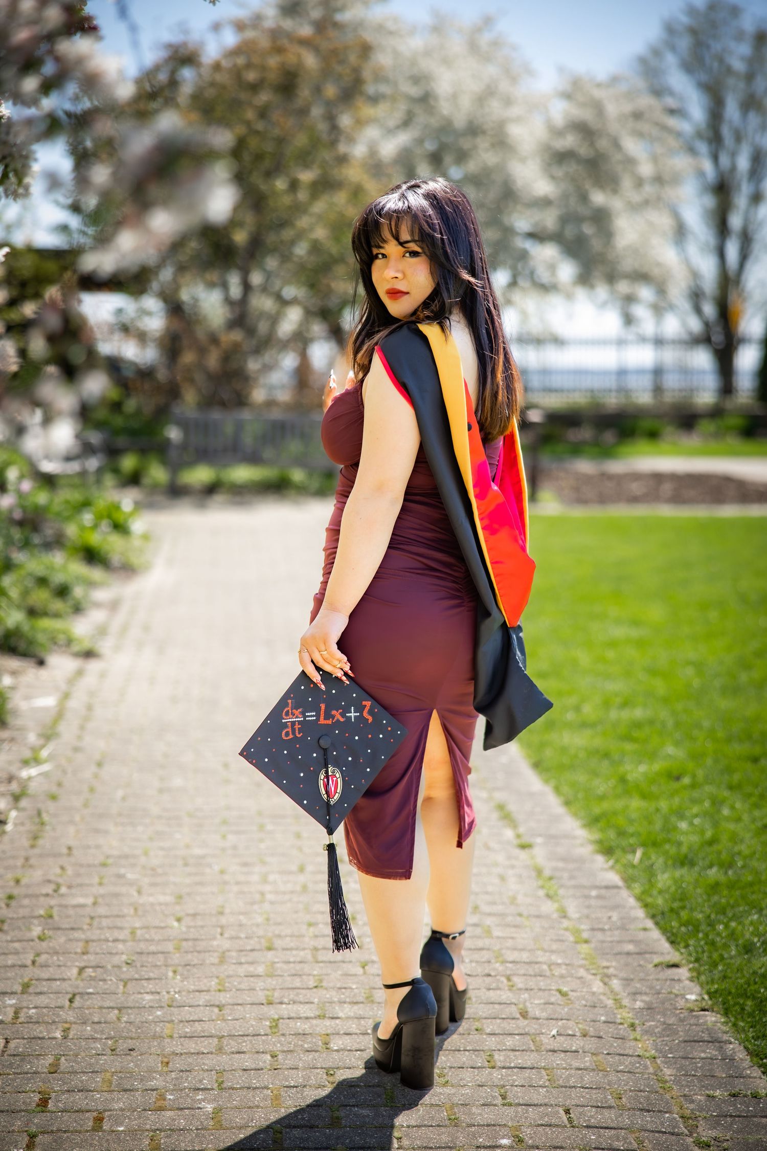 Young woman in burgundy dress holding decorated graduation cap in spring garden.