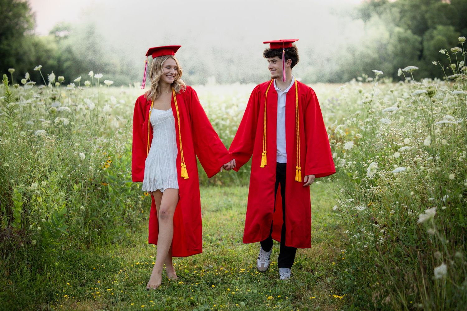Two graduates in red caps and gowns hold hands while walking through a blooming wildflower meadow.
