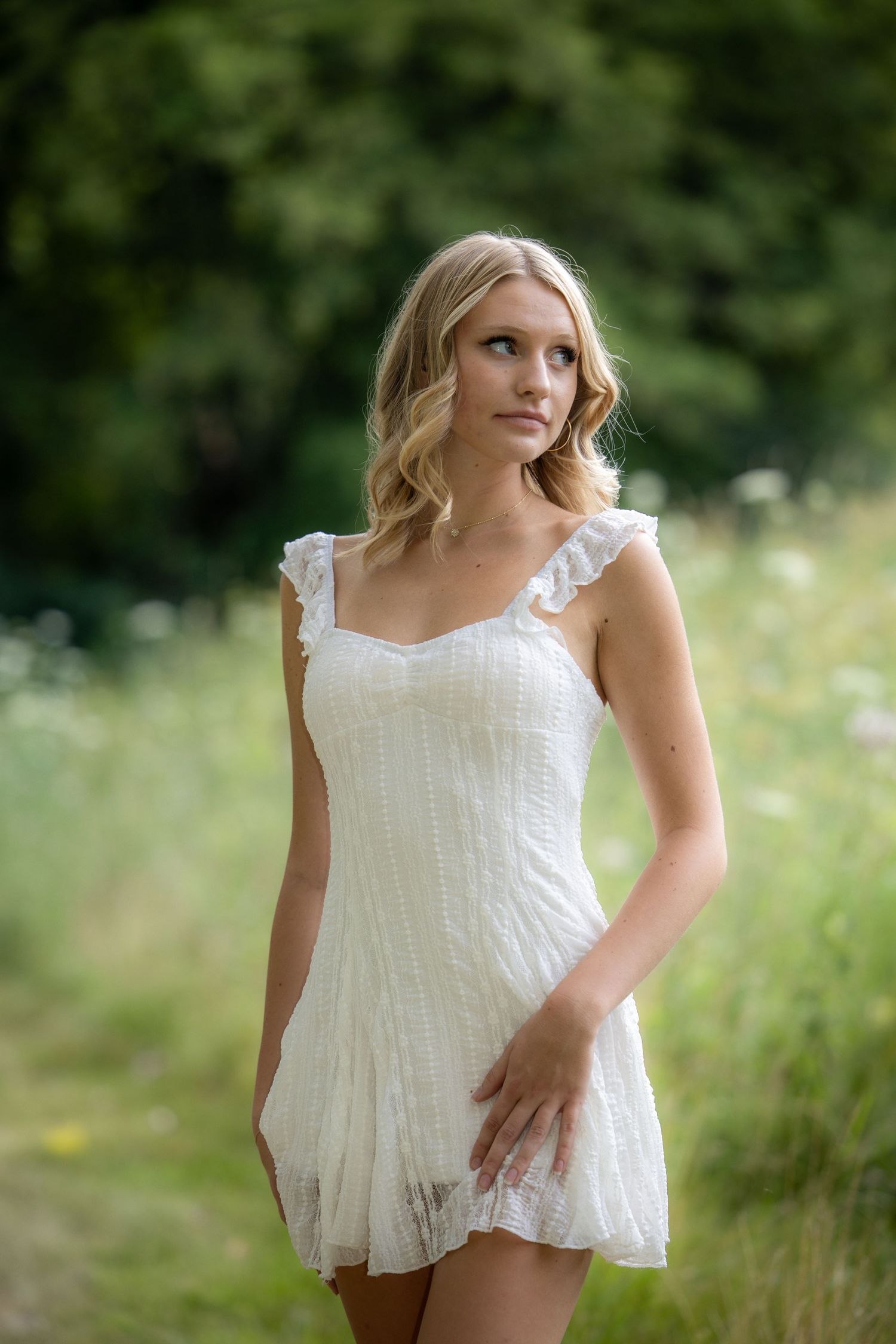 Young blonde woman in a white ruffled mini dress standing in a lush green meadow with wildflowers.
