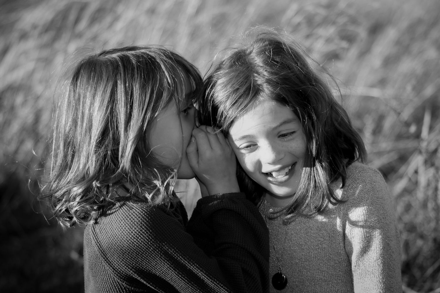 Two young girls share a secret whisper and giggle together in a charming black and white photo.