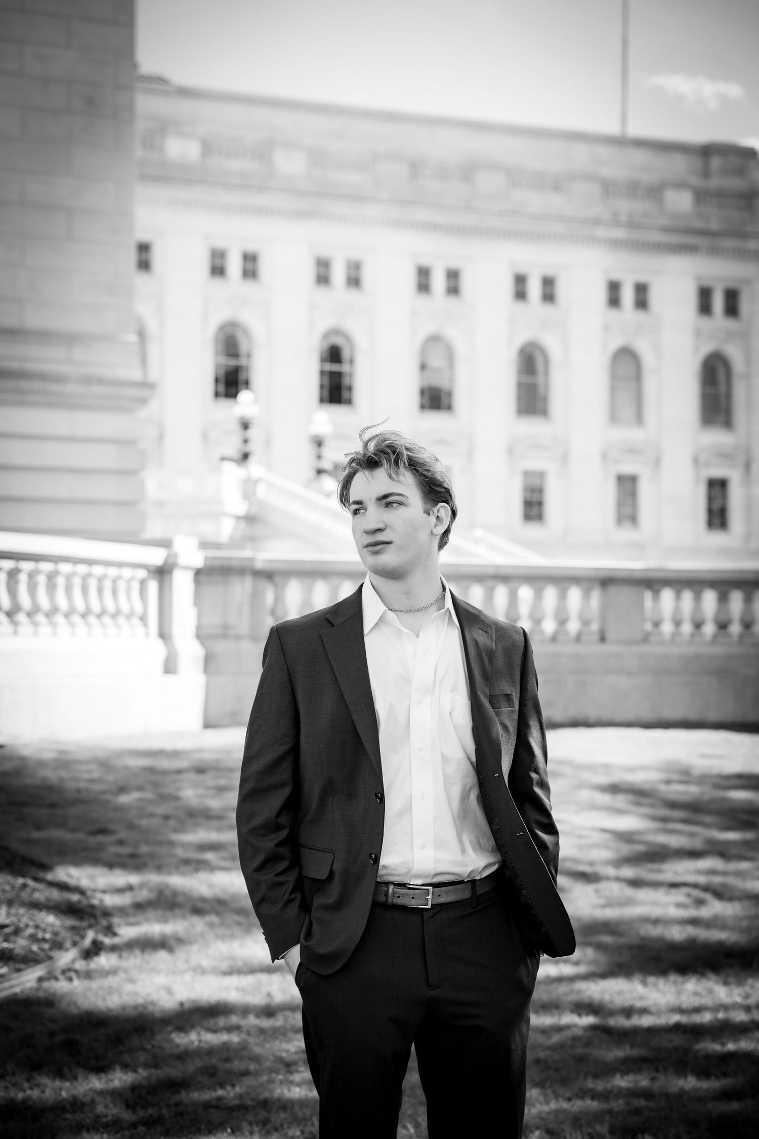 Young man in black suit poses confidently outdoors near a classical stone building in black and white photo.
