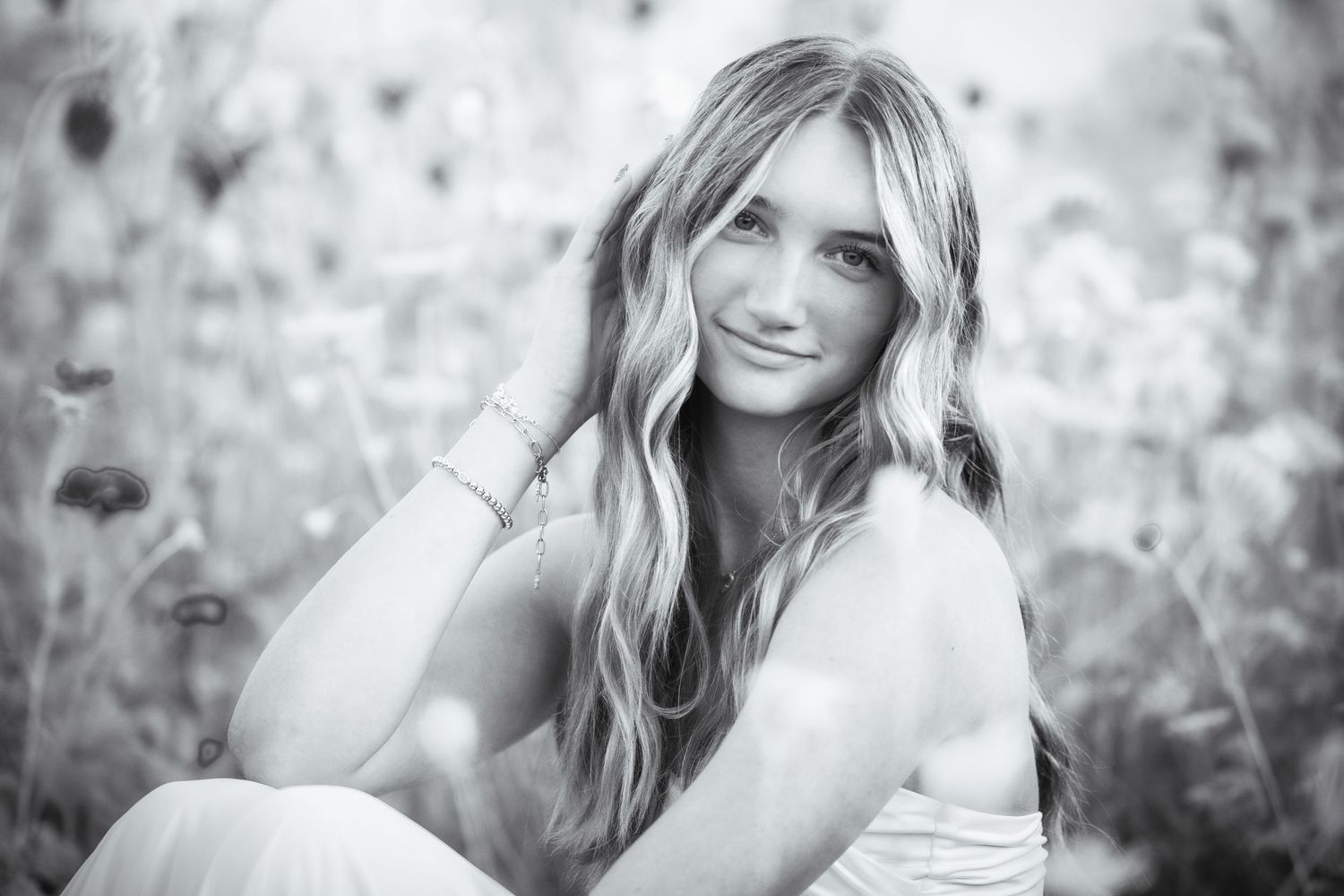 Black and white portrait of a smiling young woman with long wavy hair sitting in a wildflower field.