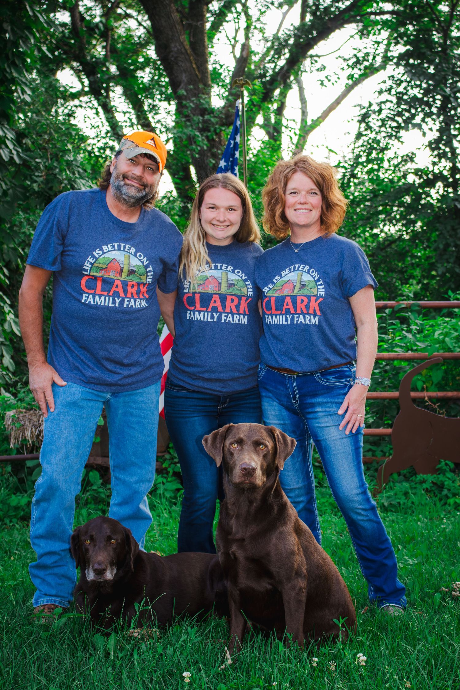 A family of three wearing Clark Family Farm shirts poses with two chocolate Labradors outdoors.