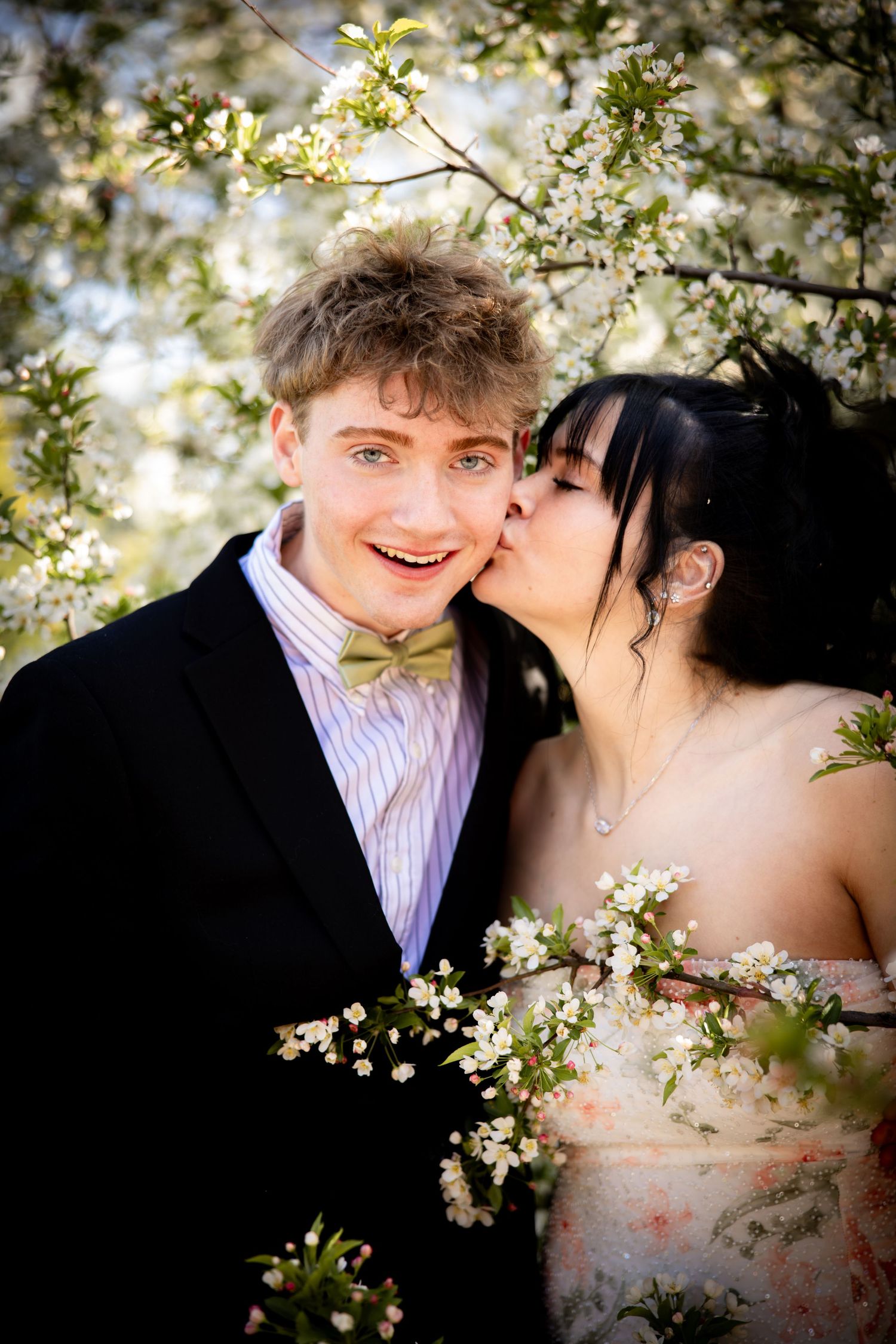 Young couple in spring cherry blossom garden, woman kissing mans cheek, man smiling in black suit with bow tie.
