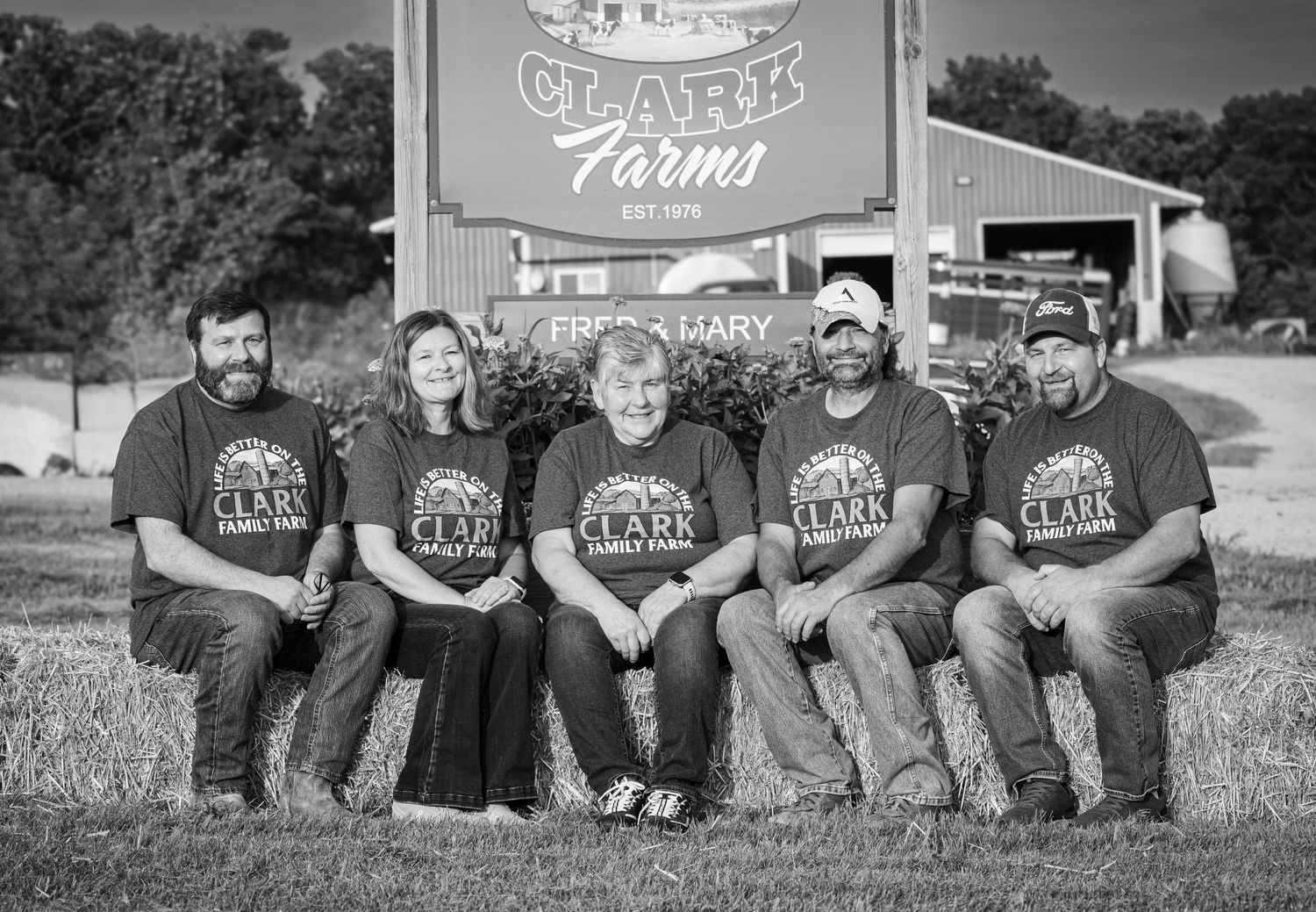 Five family members in Clark Farms shirts sit on hay bales in front of a farm sign.