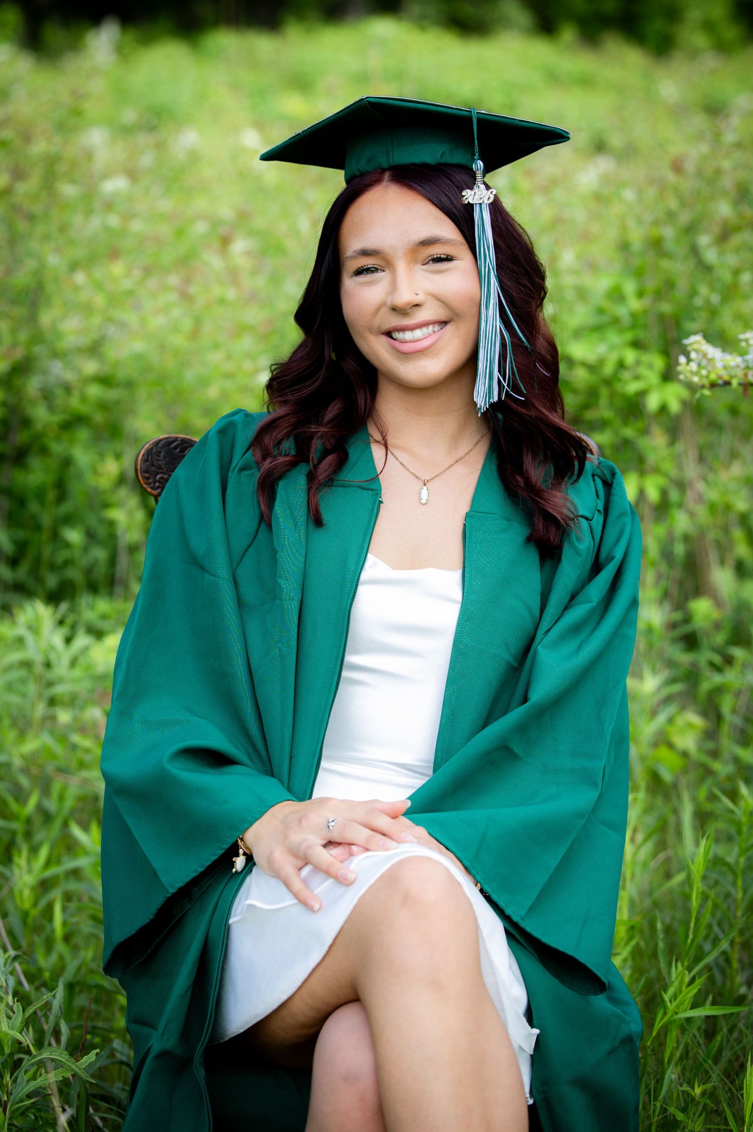 Smiling young woman in green graduation cap and gown seated in outdoor flower field.